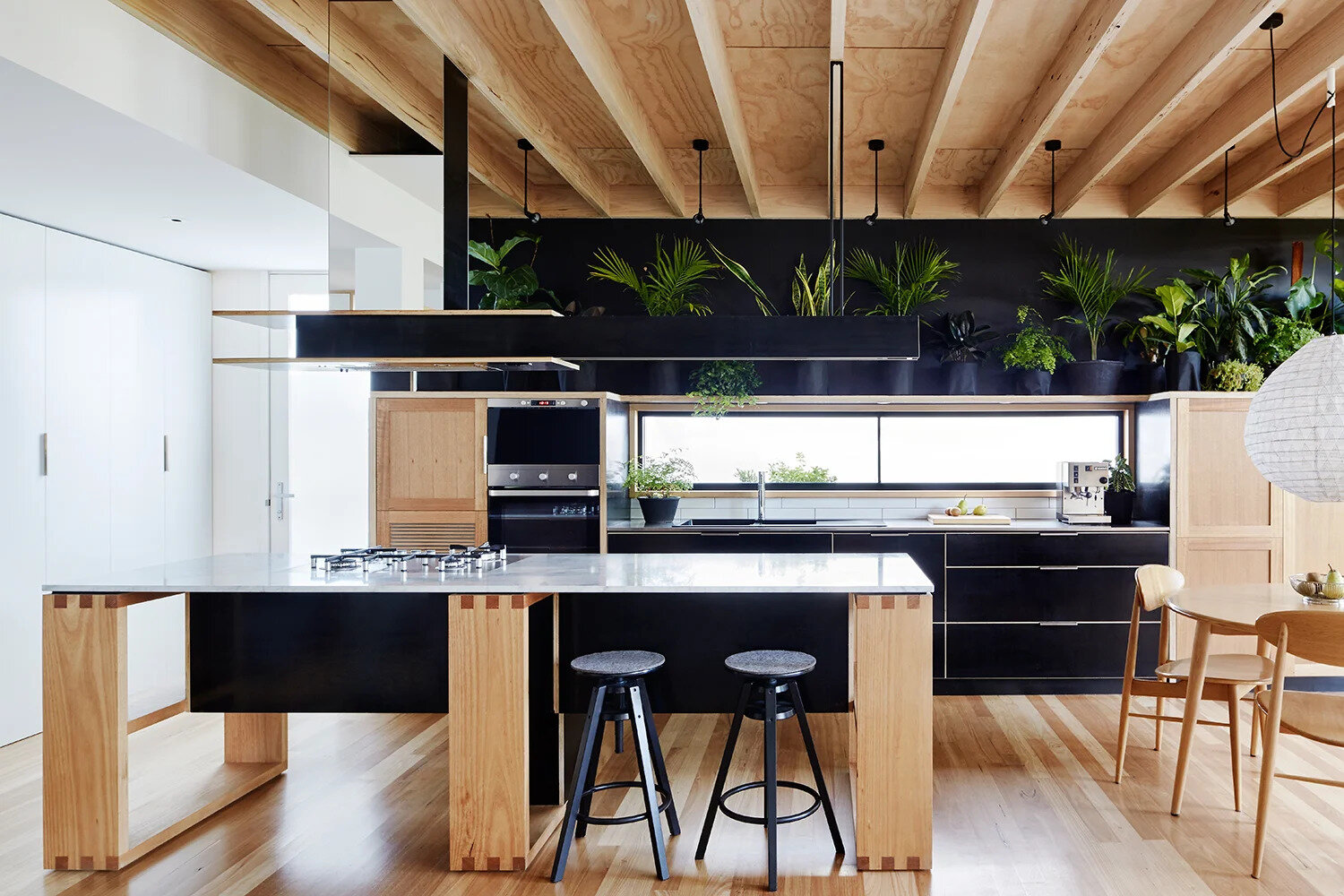 The kitchen and dining areas sit side by side, designed to feel both functional and welcoming. The kitchen pairs black formply drawer fronts with pale wood, striking a balance between boldness and warmth. A row of greenery perched above the cabinetry softens the space, adding life and texture.