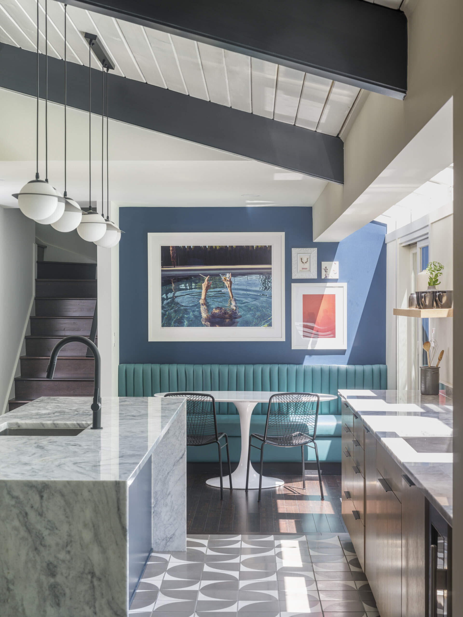 Adjacent to the kitchen, the breakfast nook centers on a custom rich blue banquette paired with a classic mid-century tulip table and wire chairs. A vibrant gallery wall backs the banquette, and floor-to-ceiling glass opens the view to the trees outside.
