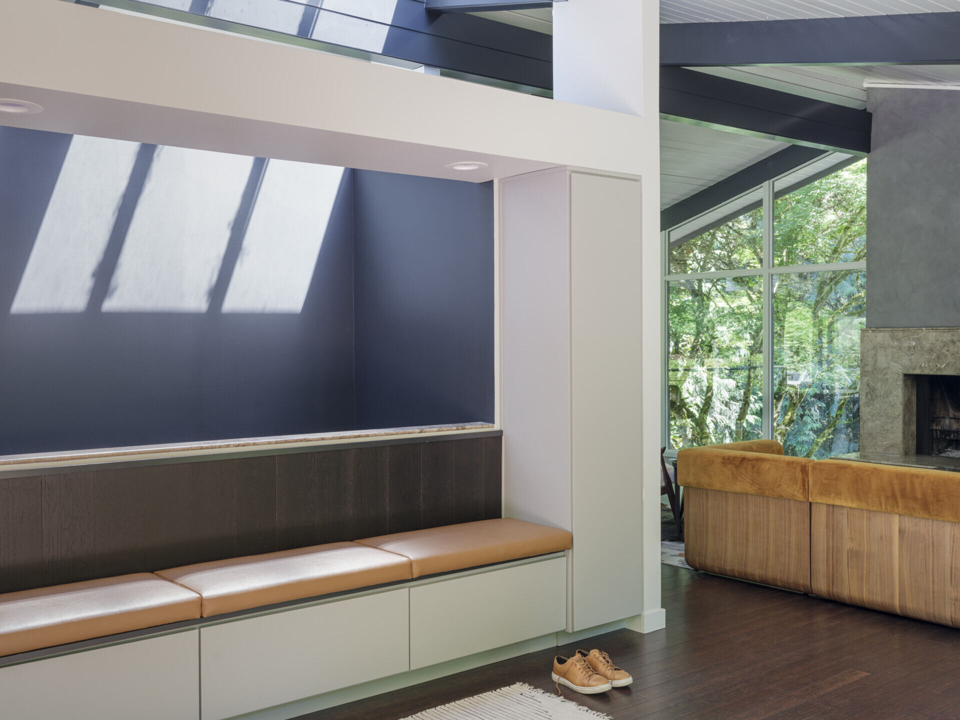 Inside, the living room leans into drama with charcoal limewash walls, a dark stone fireplace, and a burnt orange sofa that anchors the space. Surrounded by greenery through floor-to-ceiling windows, the effect is warm and grounded, balancing mid-century structure with contemporary mood.