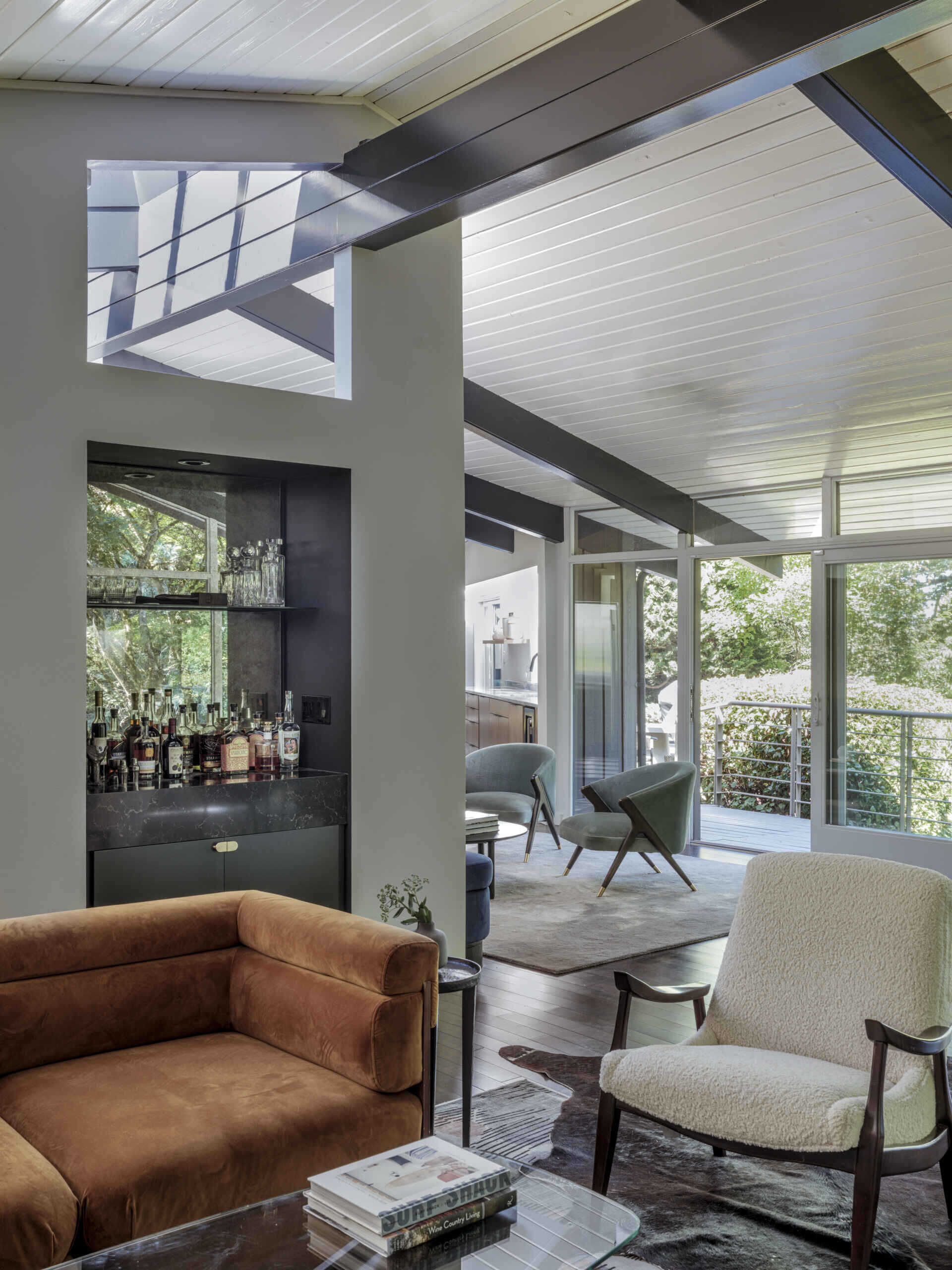 Inside, the living room leans into drama with charcoal limewash walls, a dark stone fireplace, and a burnt orange sofa that anchors the space. Surrounded by greenery through floor-to-ceiling windows, the effect is warm and grounded, balancing mid-century structure with contemporary mood.