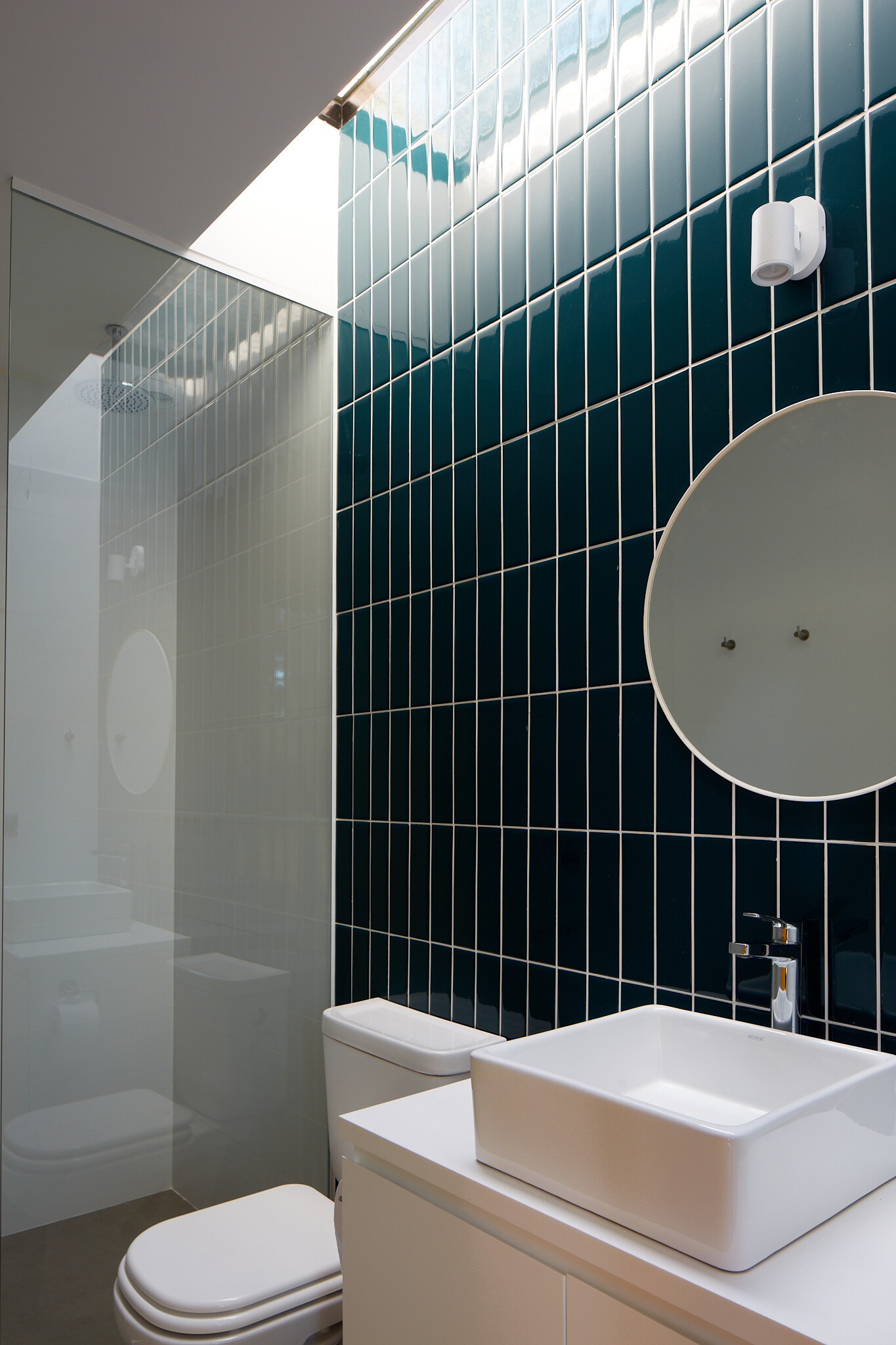 This bathroom combines simplicity with bold detail, featuring a striking wall of glossy deep teal tiles that draw the eye upward. A skylight washes the space in natural light, enhancing the contrast between the dark vertical tiles and the crisp white fixtures and shower tiles.