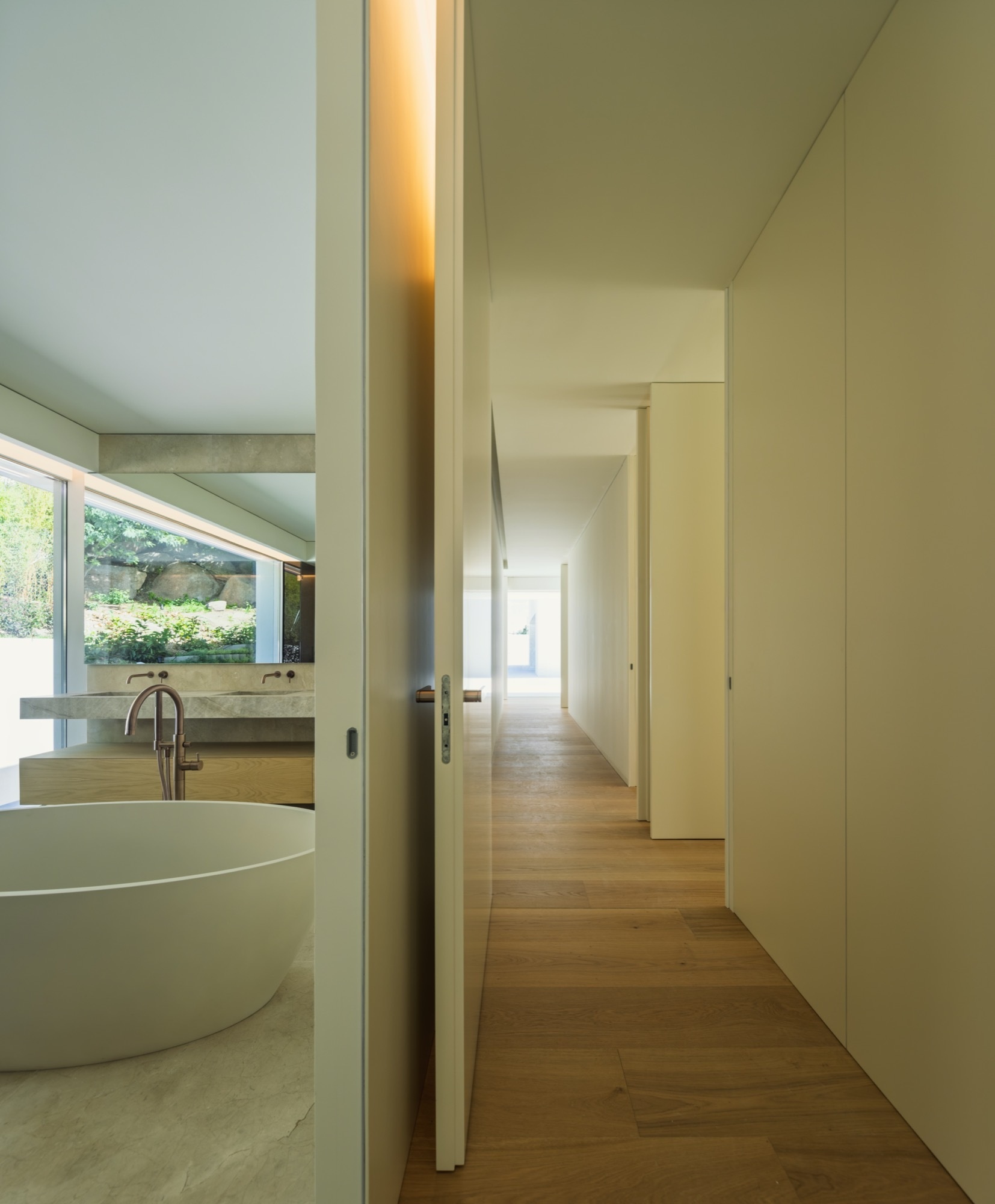 This mdoern bathroom continues the theme of warm whites and integrated materials. Ash wood, natural stone, and soft lighting temperatures help sustain the sense of calm that permeates the entire home, while a round bathtub complements the curves of the house.
