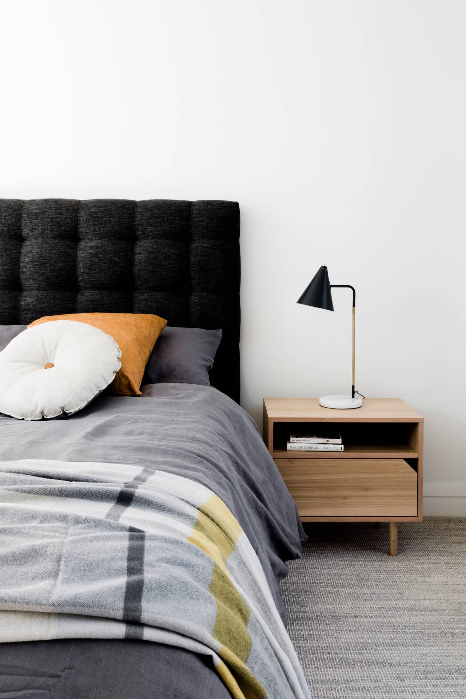 In the bedroom, restraint defines the mood. Light walls and flooring keep the space open, while a dark grey upholstered headboard and black lamp introduce contrast. A simple light wood nightstand ties back to the home’s recurring material theme, connecting the private space to the broader design language.