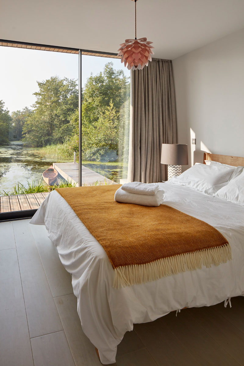A serene bedroom features floor-to-ceiling windows overlooking the calm waters of the Norfolk Broads.