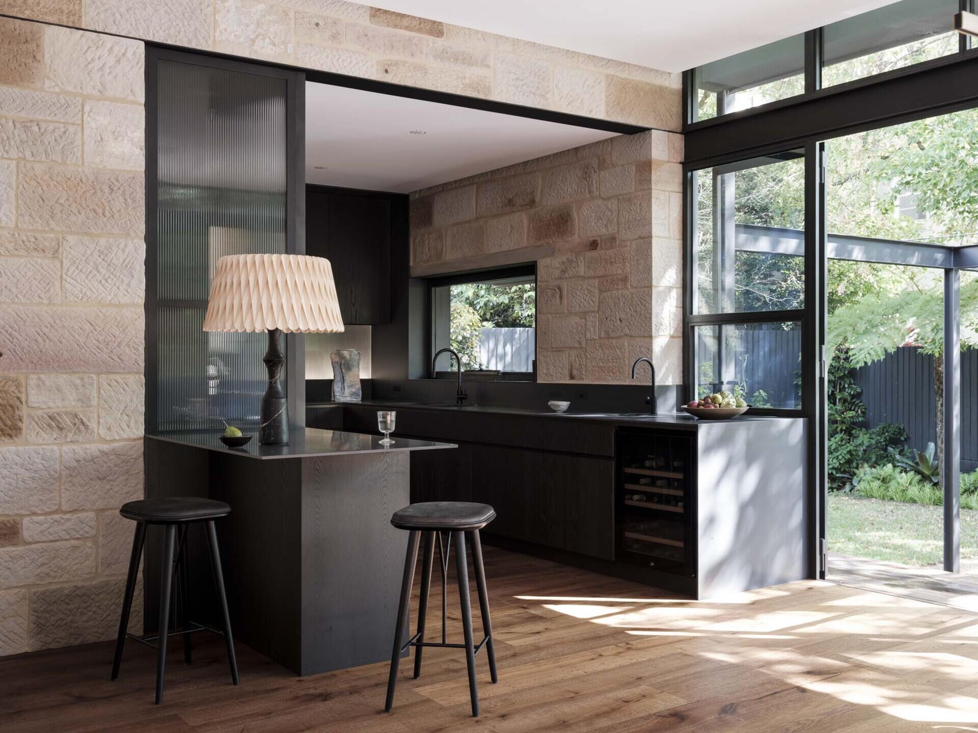 This kitchen is framed by sandstone walls that tie it back to the original cottage. Dark timber cabinetry gives the space depth, contrasted by a smooth stone benchtop that runs the length of the room. Steel-framed glazing floods the area with natural light and opens directly to the garden, while timber flooring grounds the palette in warmth.