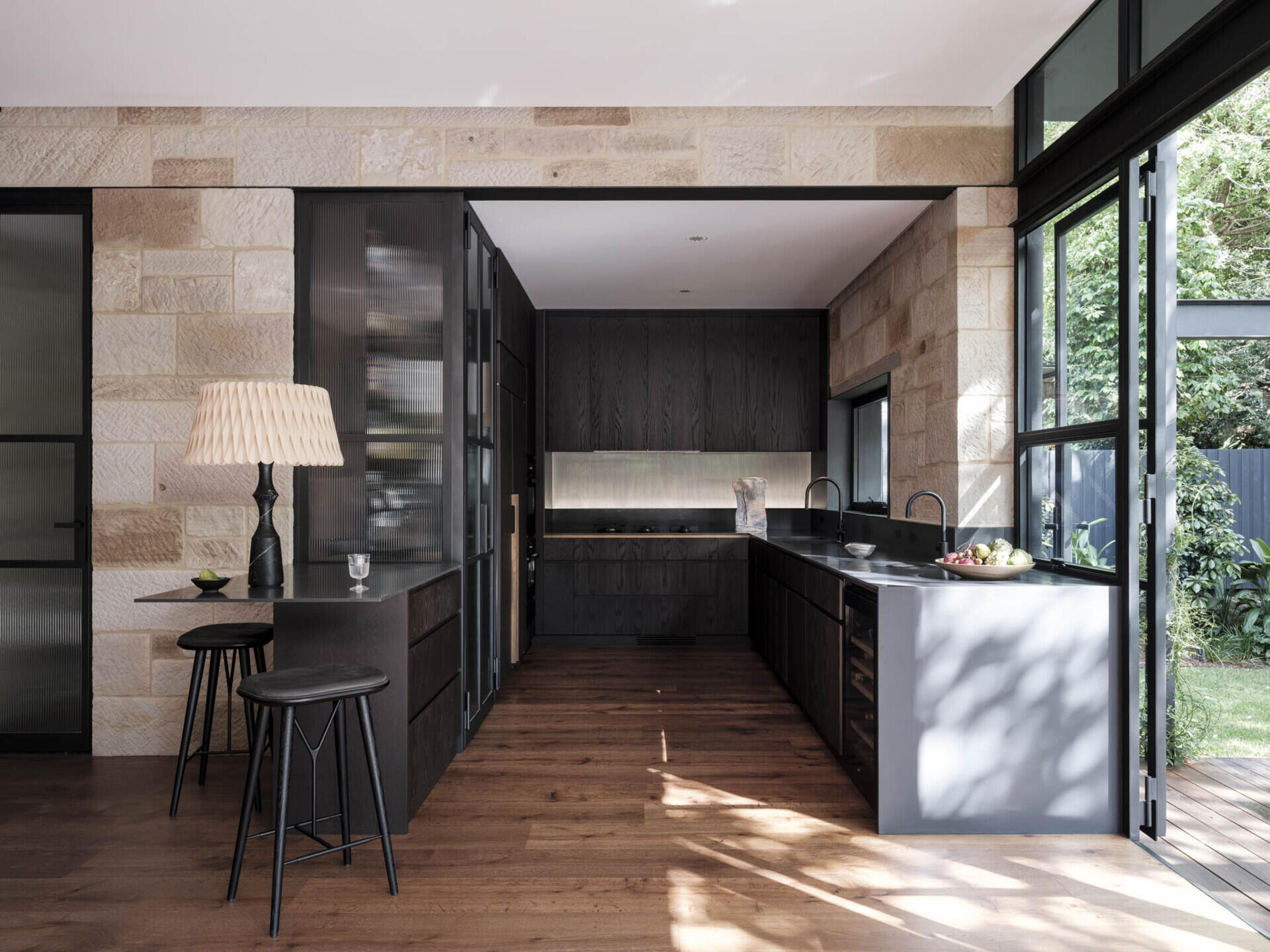 This kitchen is framed by sandstone walls that tie it back to the original cottage. Dark timber cabinetry gives the space depth, contrasted by a smooth stone benchtop that runs the length of the room. Steel-framed glazing floods the area with natural light and opens directly to the garden, while timber flooring grounds the palette in warmth.