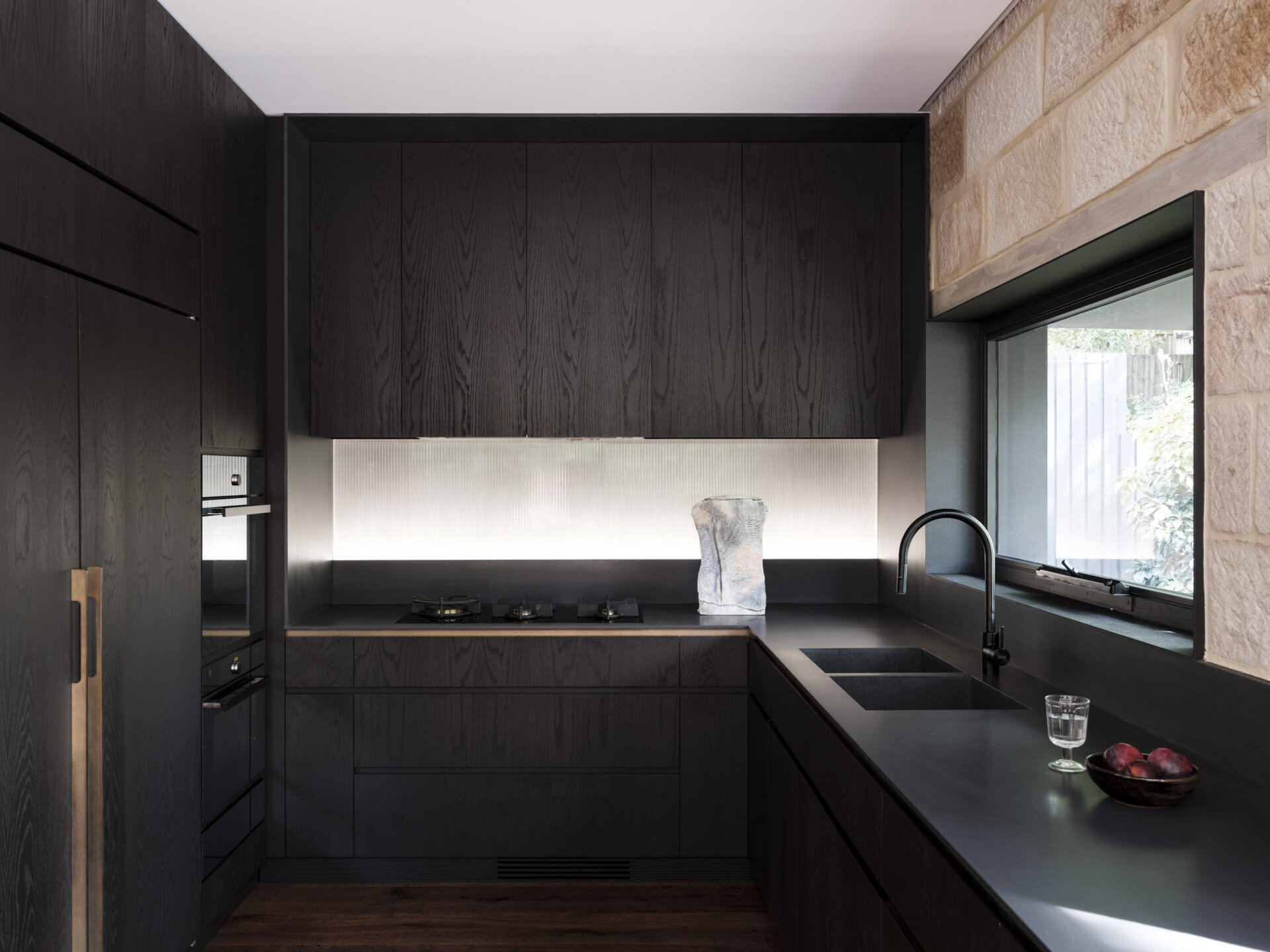 This kitchen is framed by sandstone walls that tie it back to the original cottage. Dark timber cabinetry gives the space depth, contrasted by a smooth stone benchtop that runs the length of the room. Steel-framed glazing floods the area with natural light and opens directly to the garden, while timber flooring grounds the palette in warmth.