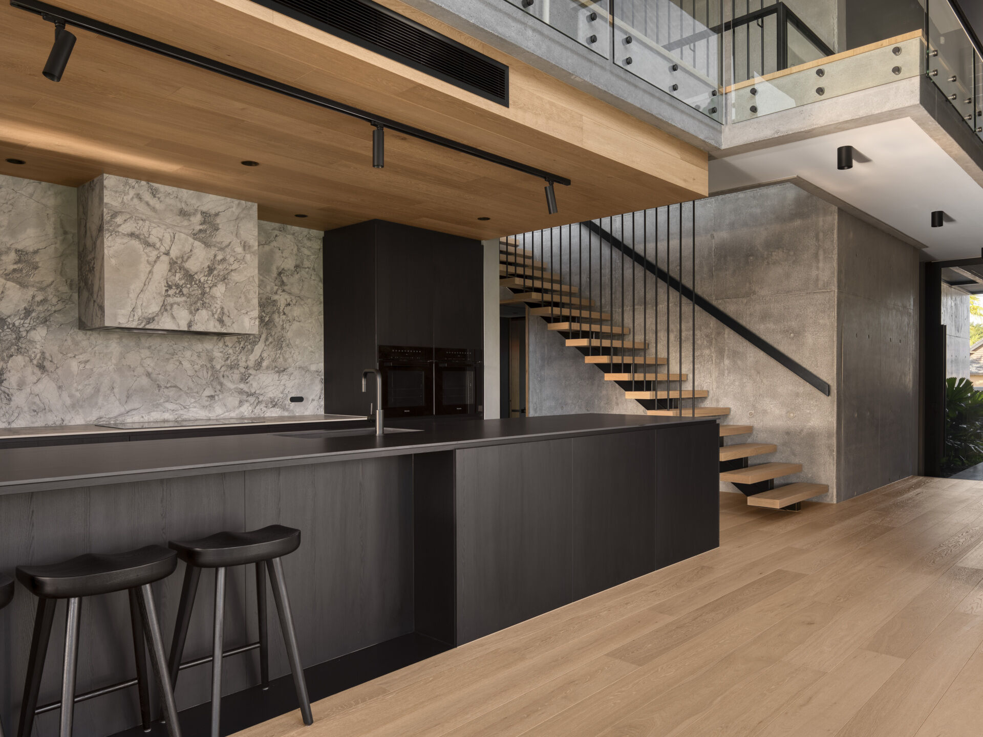 Stepping inside, the kitchen uses black-stained timber, stone, and raw concrete to combine durability with texture. Abundant natural light filters in, connecting the space to the outdoors. Adjacent, the butlers pantry supports storage and preparation, keeping the main kitchen sleek and functional. Both spaces reflect the home’s emphasis on material honesty and subtropical living.