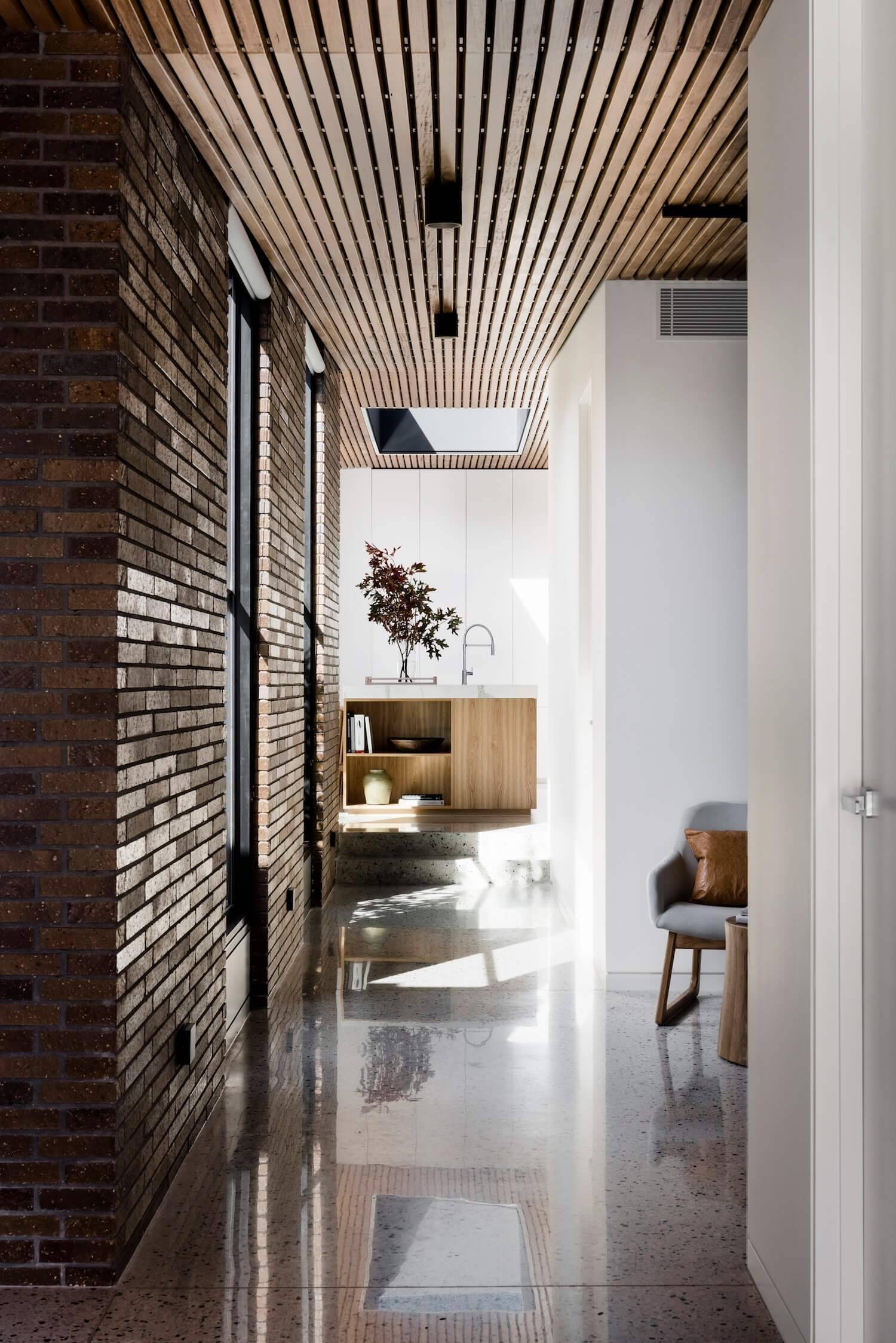 The interiors are designed with contrast in mind. A timber batten ceiling draws the eye through the space, guiding movement while warming the atmosphere. Below, white concrete flooring keeps the rooms light and airy. In the living area, a black feature wall defines the space within the open-plan layout, balancing the softer tones of wood and brick.