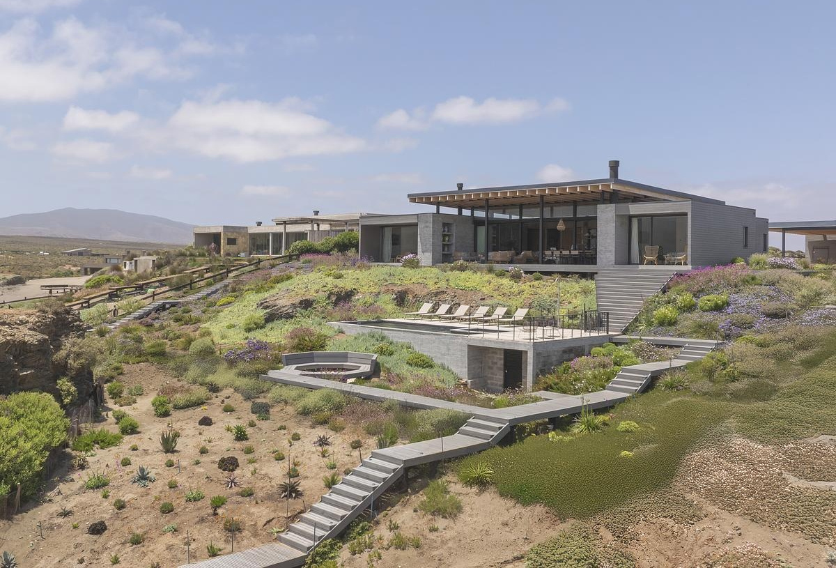 Terraced paths of timber decking guide you down through native plantings, where succulents, grasses, and flowering shrubs stitch the house into the coastal terrain. At the center of it all sits the pool, carved into a lower terrace with a sun loungers facing the horizon. A fire pit nearby adds another gathering spot, grounding evenings in warmth as the coastline stretches away.