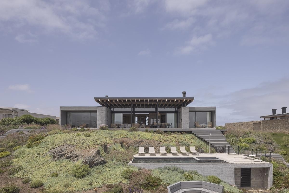 Terraced paths of timber decking guide you down through native plantings, where succulents, grasses, and flowering shrubs stitch the house into the coastal terrain. At the center of it all sits the pool, carved into a lower terrace with a sun loungers facing the horizon. A fire pit nearby adds another gathering spot, grounding evenings in warmth as the coastline stretches away.
