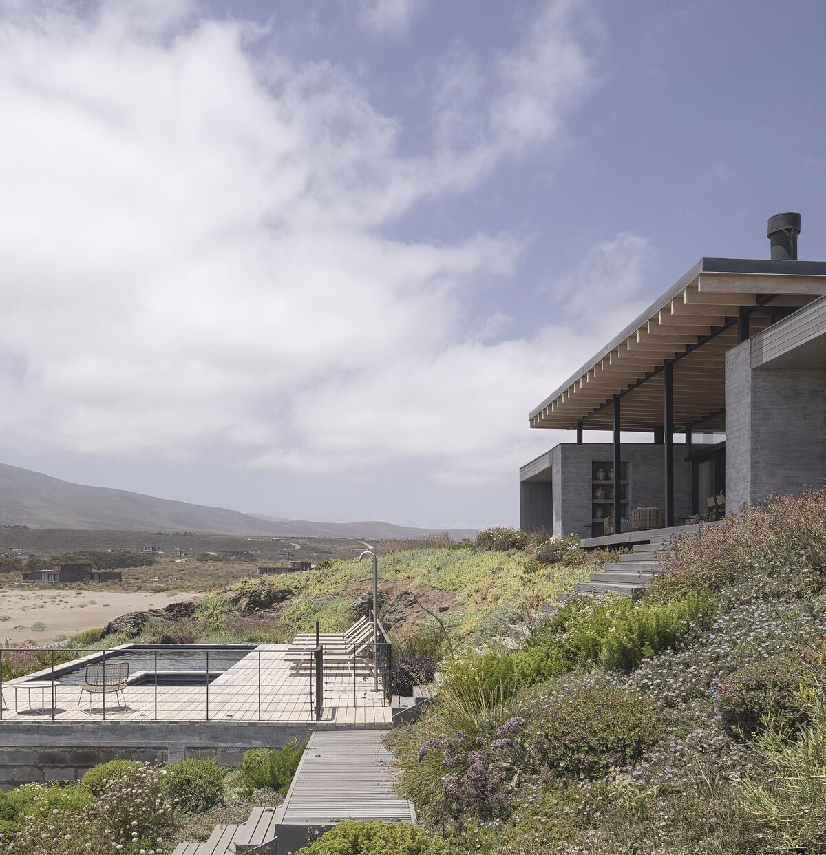 Terraced paths of timber decking guide you down through native plantings, where succulents, grasses, and flowering shrubs stitch the house into the coastal terrain. At the center of it all sits the pool, carved into a lower terrace with a sun loungers facing the horizon. A fire pit nearby adds another gathering spot, grounding evenings in warmth as the coastline stretches away.