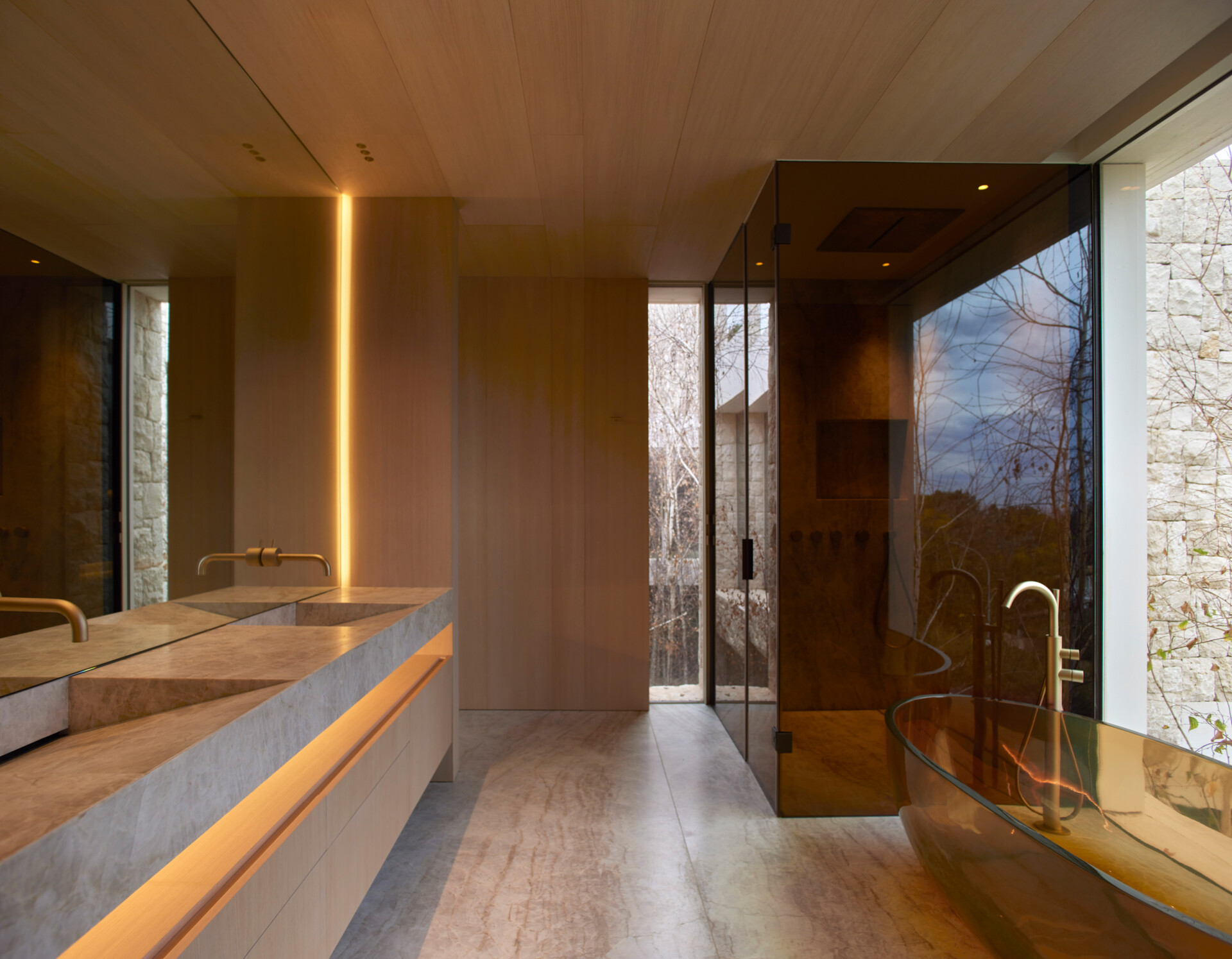 In this bathroom, stone and wood surfaces set a calm, grounding tone, while soft lighting built into the walls adds warmth and depth. A sculptural glass bathtub and matching glass-enclosed shower anchors the space, paired with sleek brass fixtures that elevate the design.