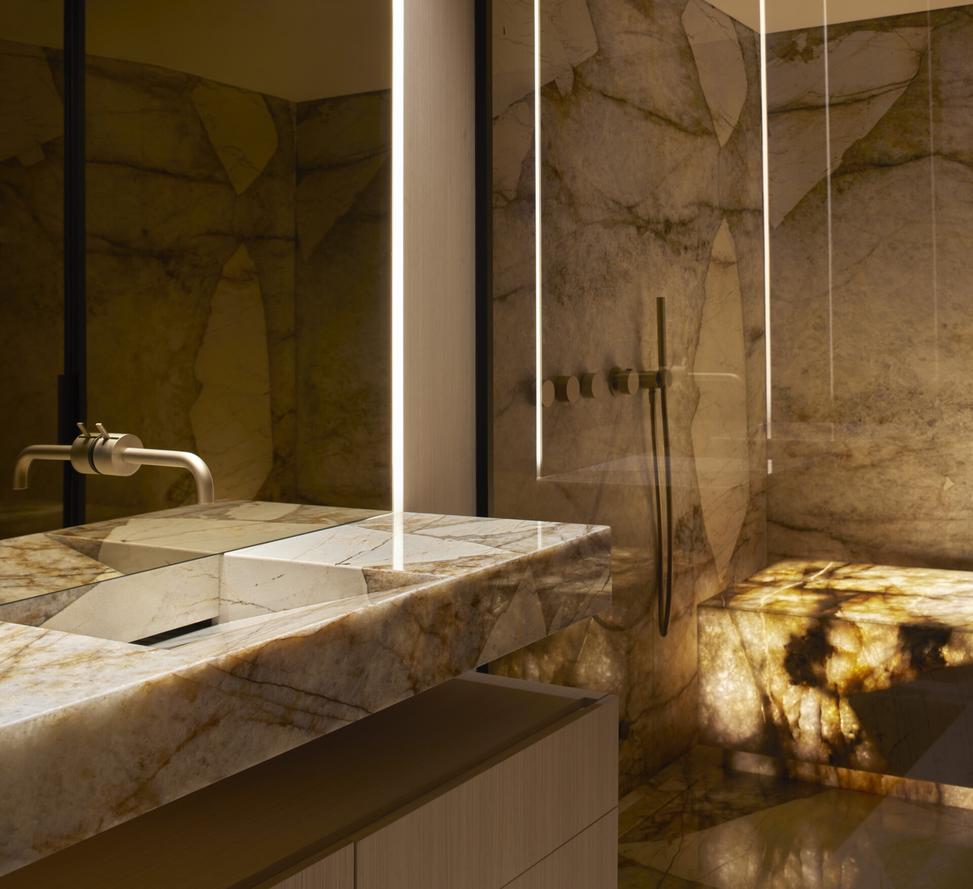 This bathroom takes on a dramatic character, wrapped in richly veined stone that makes the entire space feel sculpted from a single block. Warm backlighting highlights the textures, casting a soft glow across the walls and shower. A stone vanity with sharp geometric lines pairs with minimalist fixtures in brushed metal, keeping the focus on the natural material.