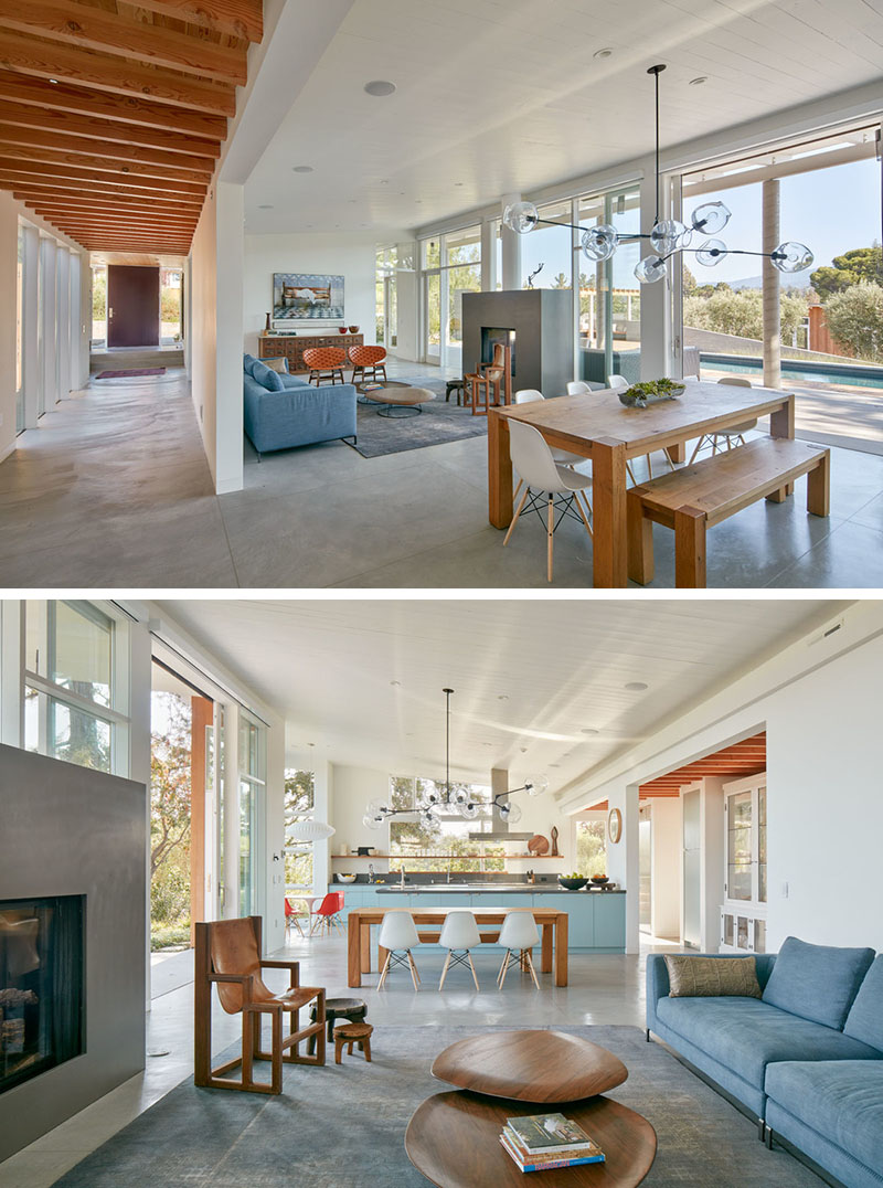 The living area features a double-sided fireplace, warming both the living room and the outdoor lounge. A sculptural pendant light hangs over a long wood dining table that anchors the space. Large sliding glass doors open the interior to the pool deck, blending indoor and outdoor living.