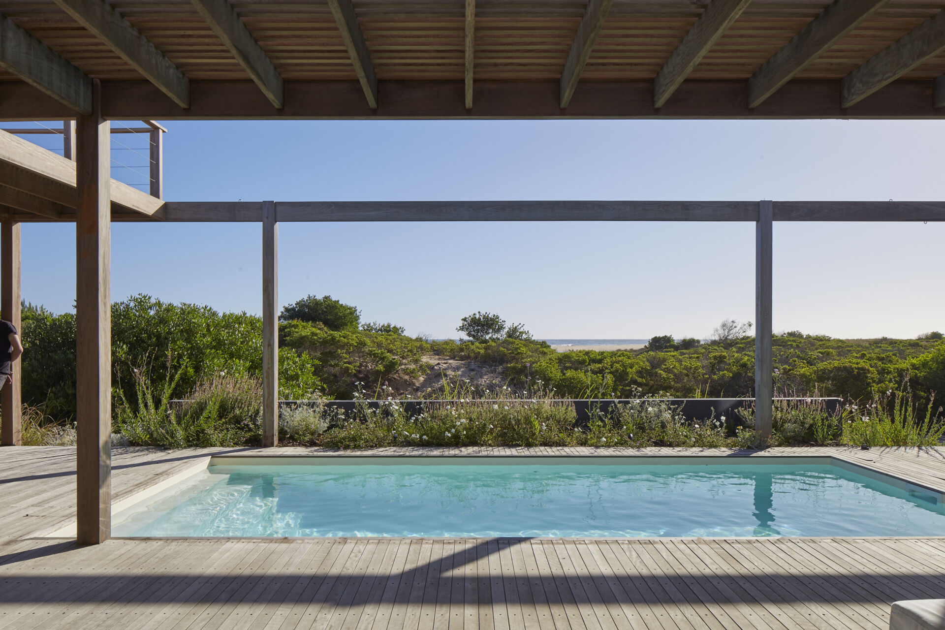 This prefabricated modular house, clad in wood, has a swimming pool, and preserves the sandy terrain while opening all rooms toward the sea.