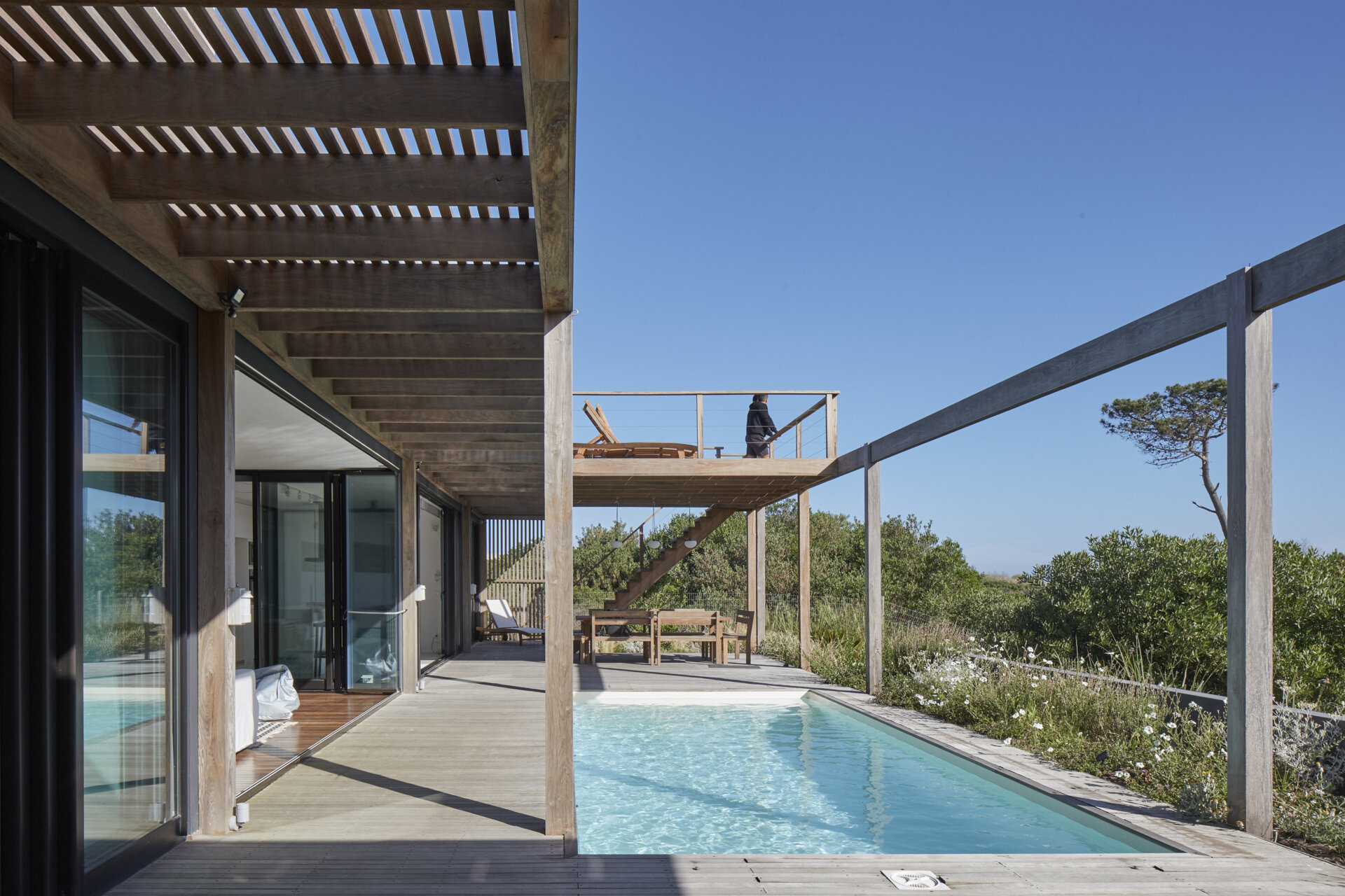 This prefabricated modular house, clad in wood, has a swimming pool, and preserves the sandy terrain while opening all rooms toward the sea.