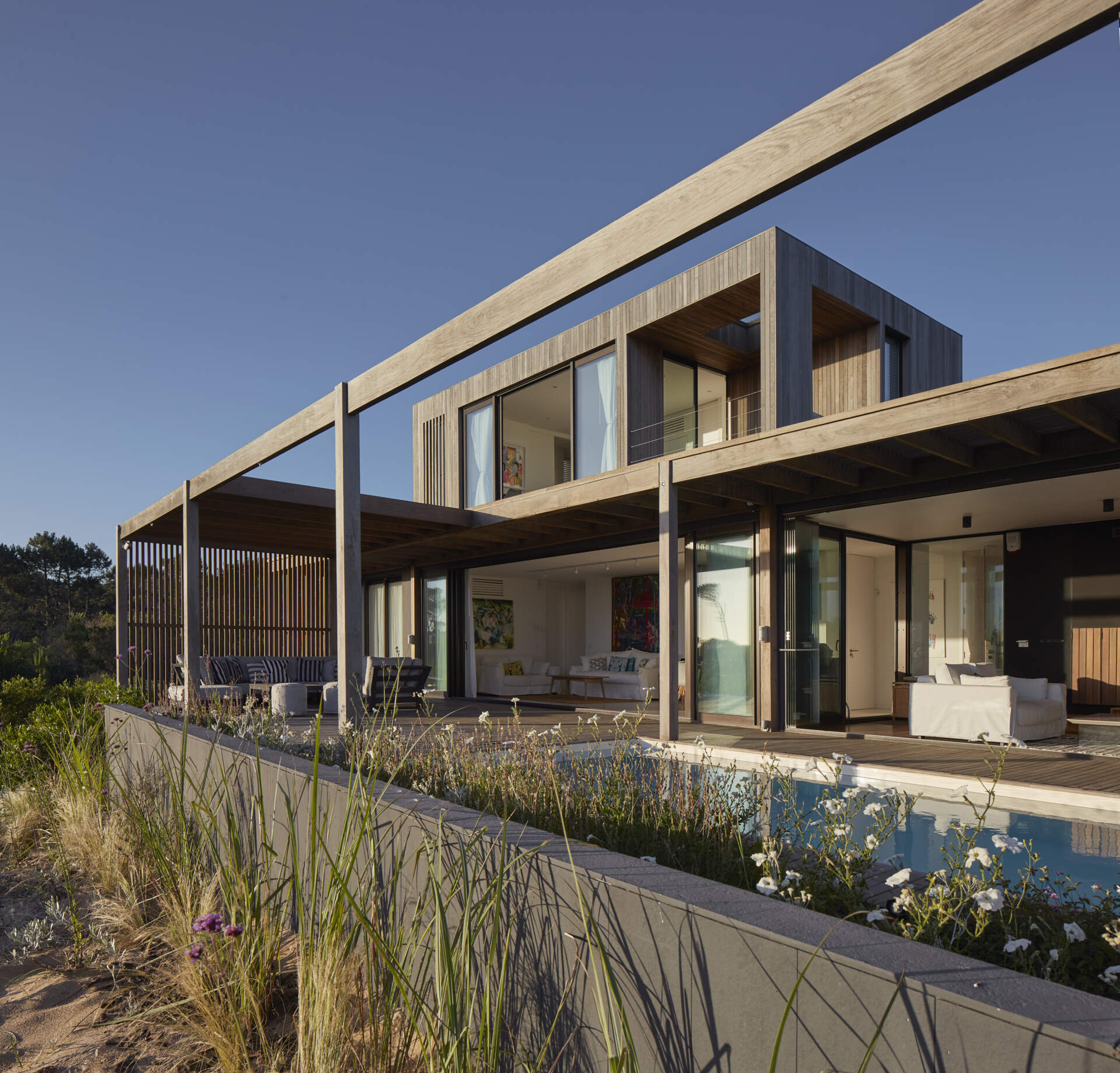 This prefabricated modular house, clad in wood, has a swimming pool, and preserves the sandy terrain while opening all rooms toward the sea.