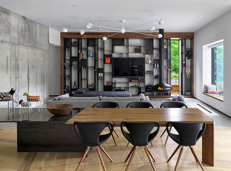 At the heart of the home is a long dining table that organizes the open-plan layout. On one side sits a family room featuring a custom-designed metal bookcase and a dramatic suspended black wood-burning fireplace.