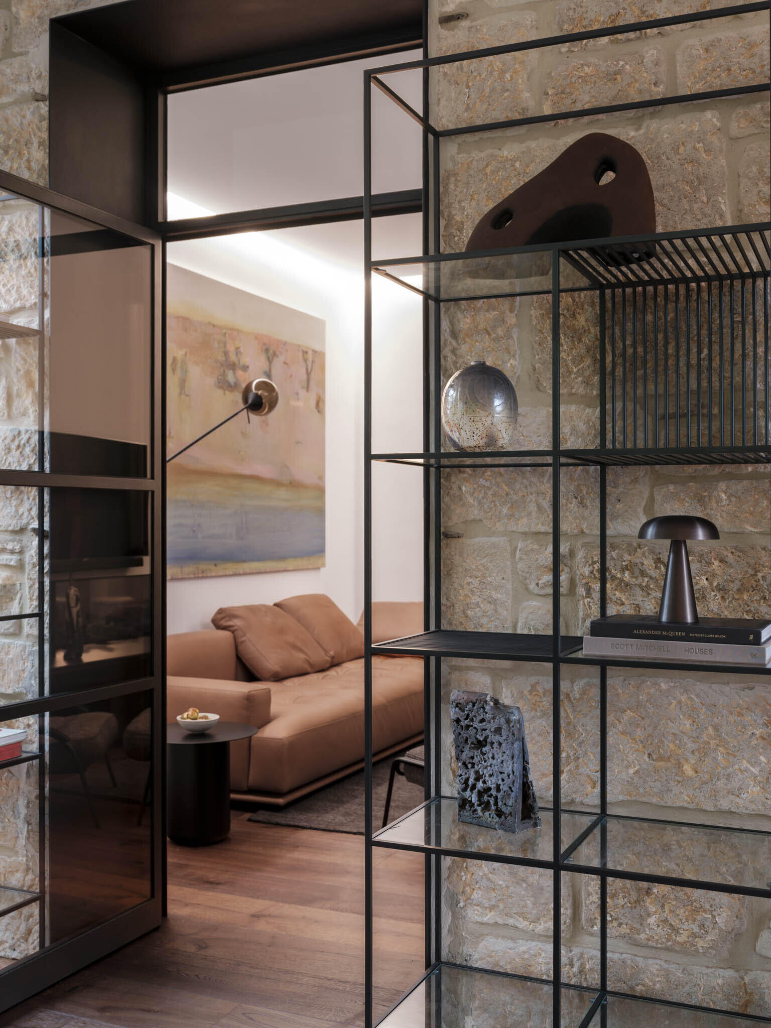 The living room, accessed via the study or the hallway, pairs restraint with comfort, where a low-slung tan sofa anchors the space against crisp white walls. Above, an extended black wall light cuts across the room like a sculptural gesture, balancing function with bold form.