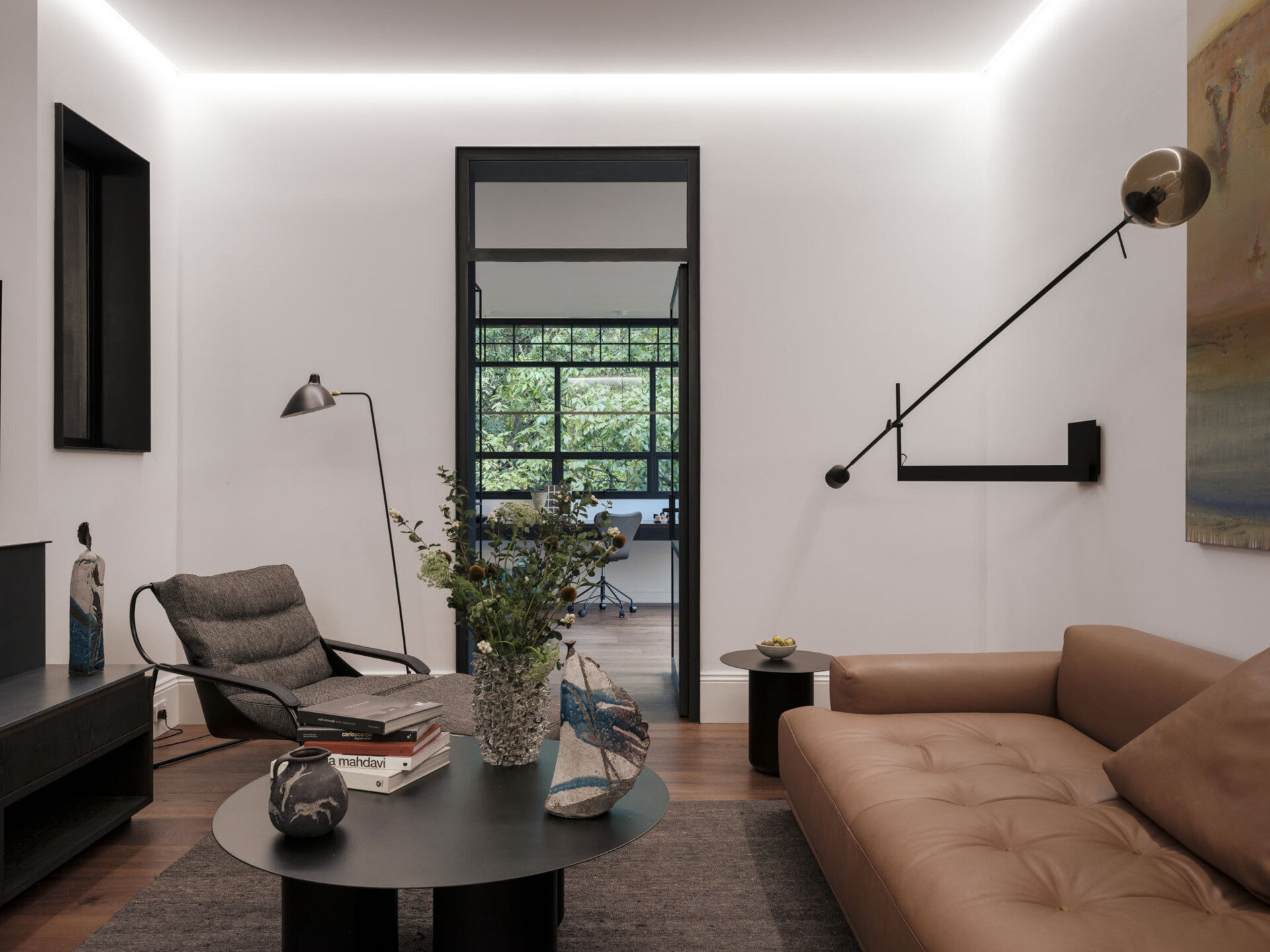 The living room, accessed via the study or the hallway, pairs restraint with comfort, where a low-slung tan sofa anchors the space against crisp white walls. Above, an extended black wall light cuts across the room like a sculptural gesture, balancing function with bold form.