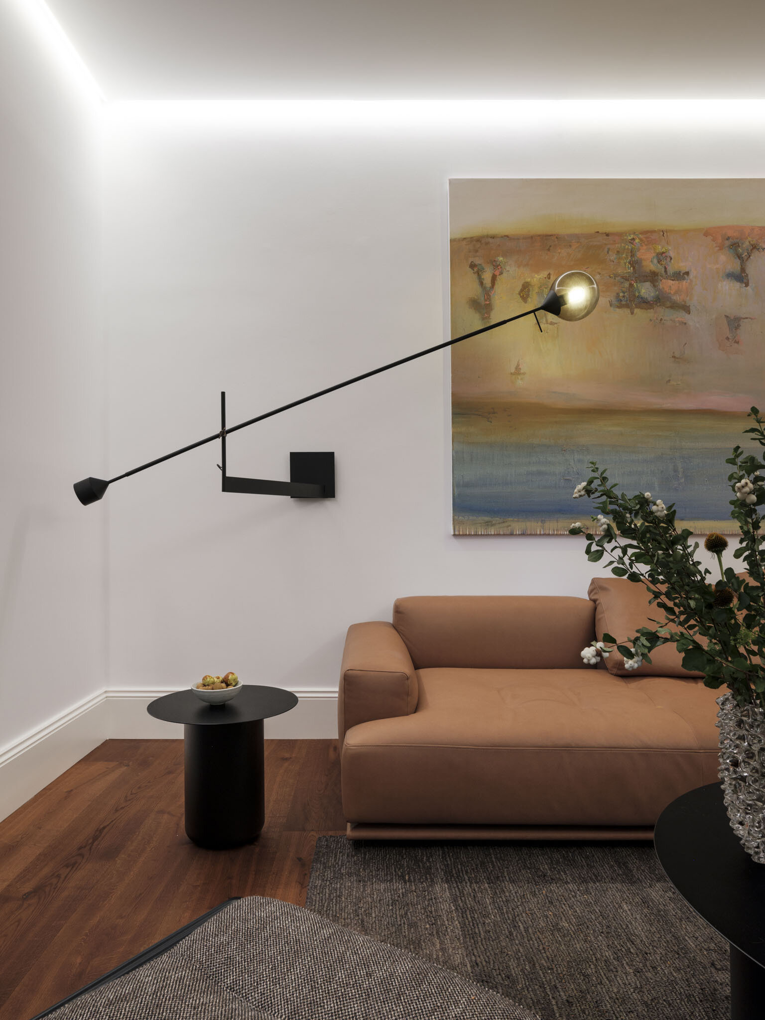 The living room, accessed via the study or the hallway, pairs restraint with comfort, where a low-slung tan sofa anchors the space against crisp white walls. Above, an extended black wall light cuts across the room like a sculptural gesture, balancing function with bold form.