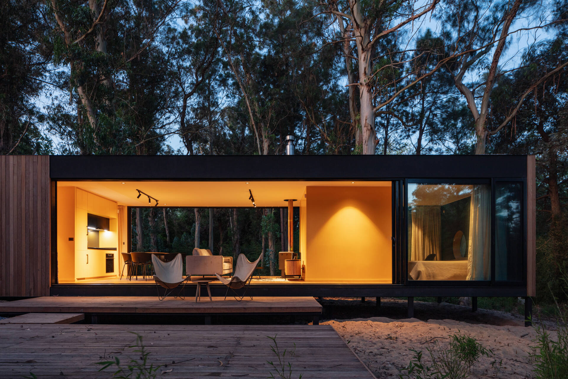 At night, the house glows warmly against the dark forest backdrop, its open-plan design fully visible through wide glass walls. The golden interior lighting creates a striking contrast with the surrounding trees, making the structure appear like a lantern tucked among the woods.