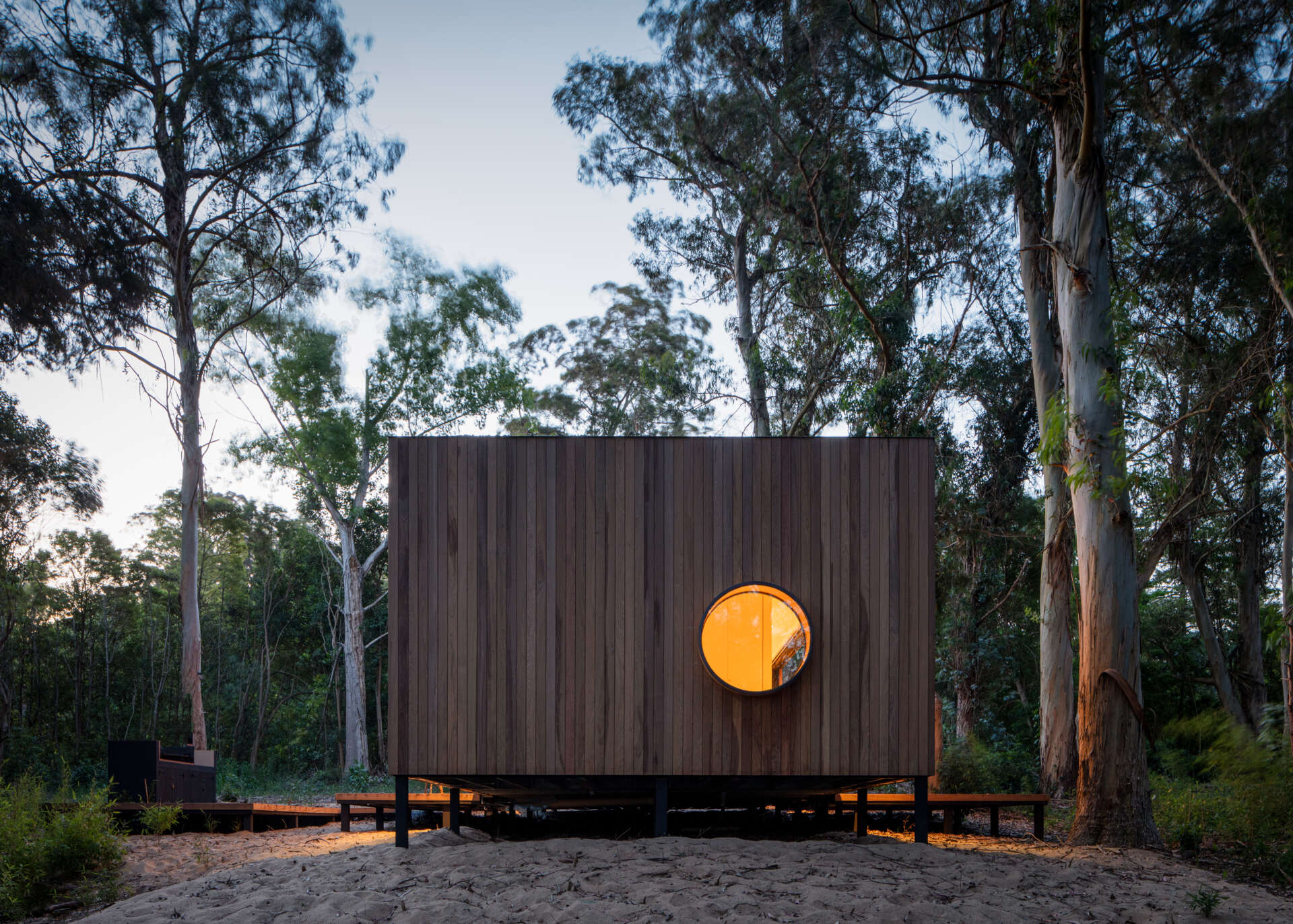 At night, the house glows warmly against the dark forest backdrop, its open-plan design fully visible through wide glass walls. The golden interior lighting creates a striking contrast with the surrounding trees, making the structure appear like a lantern tucked among the woods.