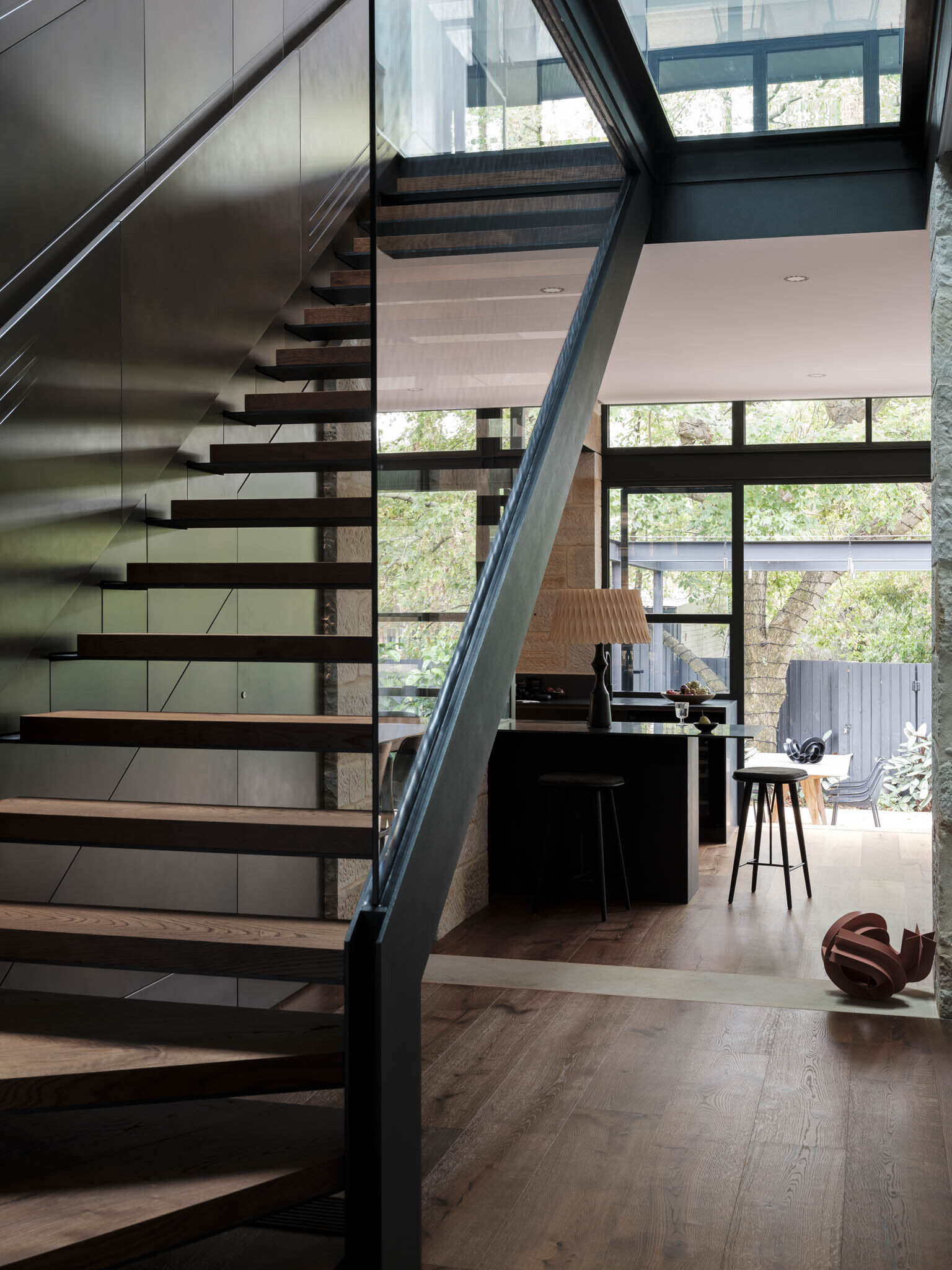 This kitchen is framed by sandstone walls that tie it back to the original cottage. Dark timber cabinetry gives the space depth, contrasted by a smooth stone benchtop that runs the length of the room. Steel-framed glazing floods the area with natural light and opens directly to the garden, while timber flooring grounds the palette in warmth.