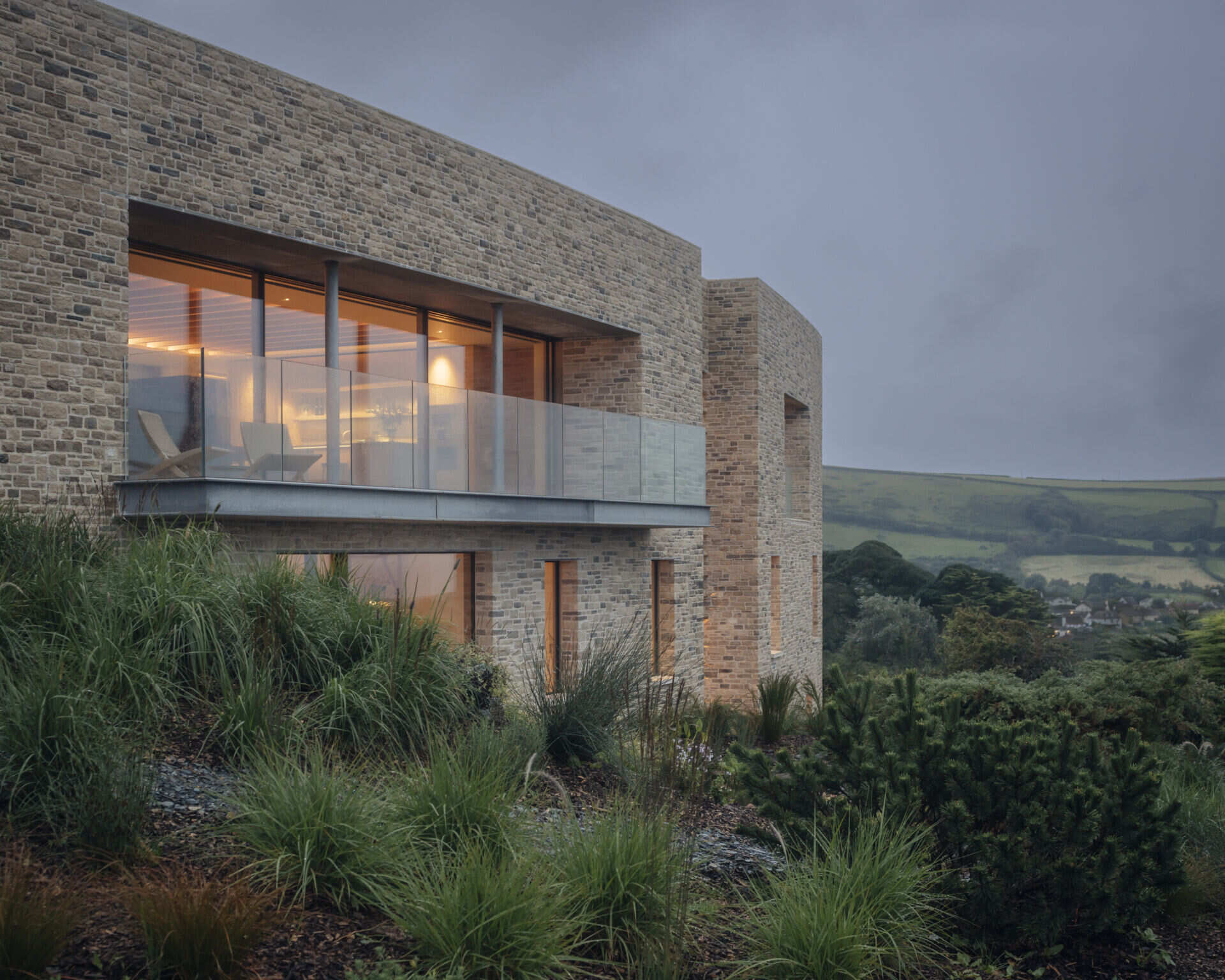 This modern home is built from pale Purbeck stone, with a curved facade that follows the natural bend of the site. A green roof hides solar panels, while coastal grasses and granite landscaping root the home into the hillside.