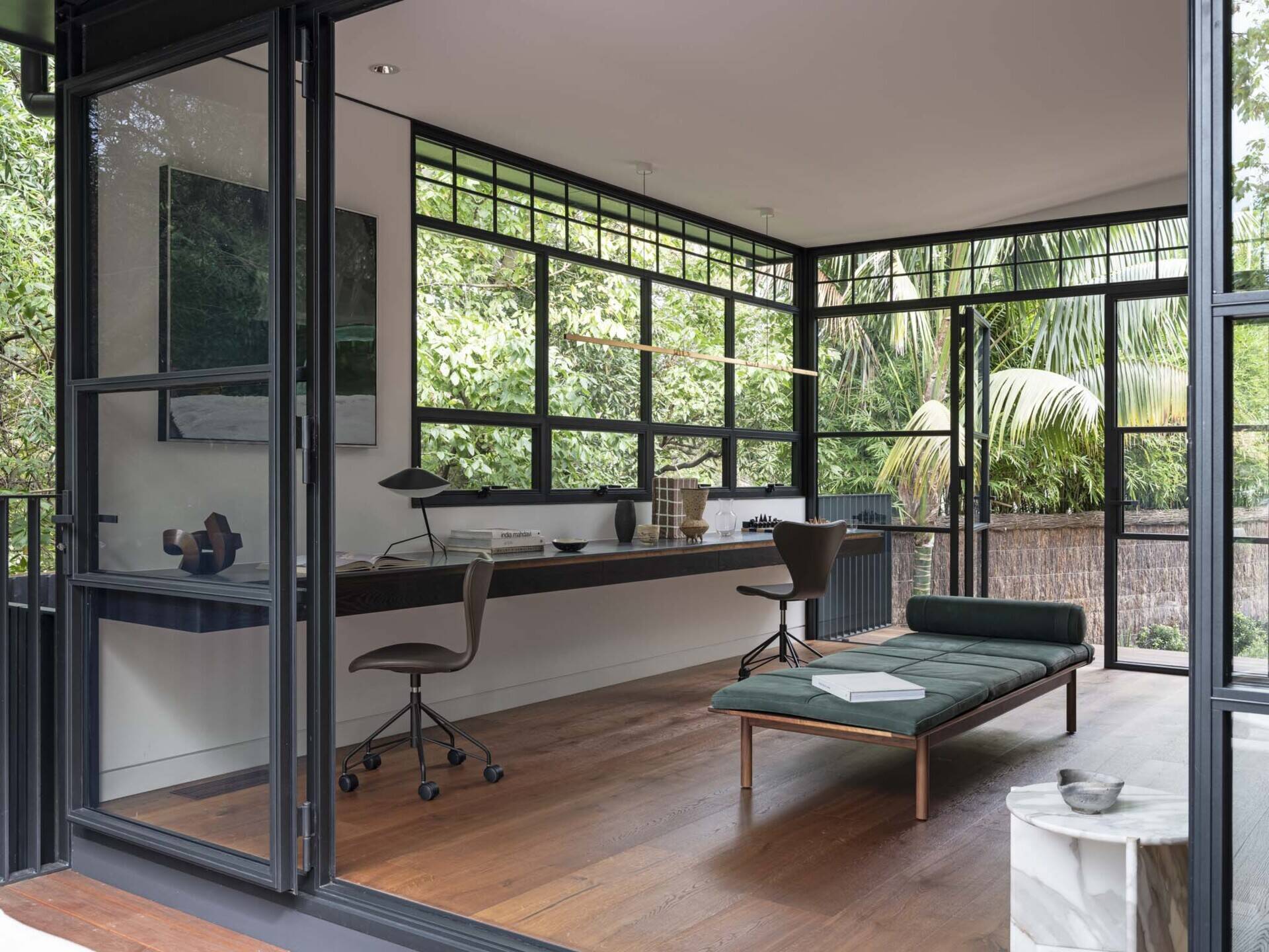 The study is wrapped in steel-framed glazing that dissolves the boundary between indoors and the surrounding greenery. A long desk stretches across one wall, offering an uninterrupted workspace anchored by minimalist chairs.