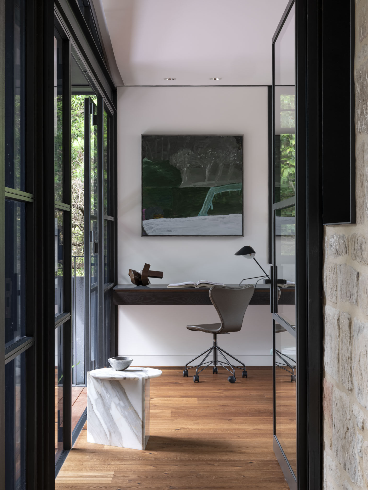 The study is wrapped in steel-framed glazing that dissolves the boundary between indoors and the surrounding greenery. A long desk stretches across one wall, offering an uninterrupted workspace anchored by minimalist chairs.