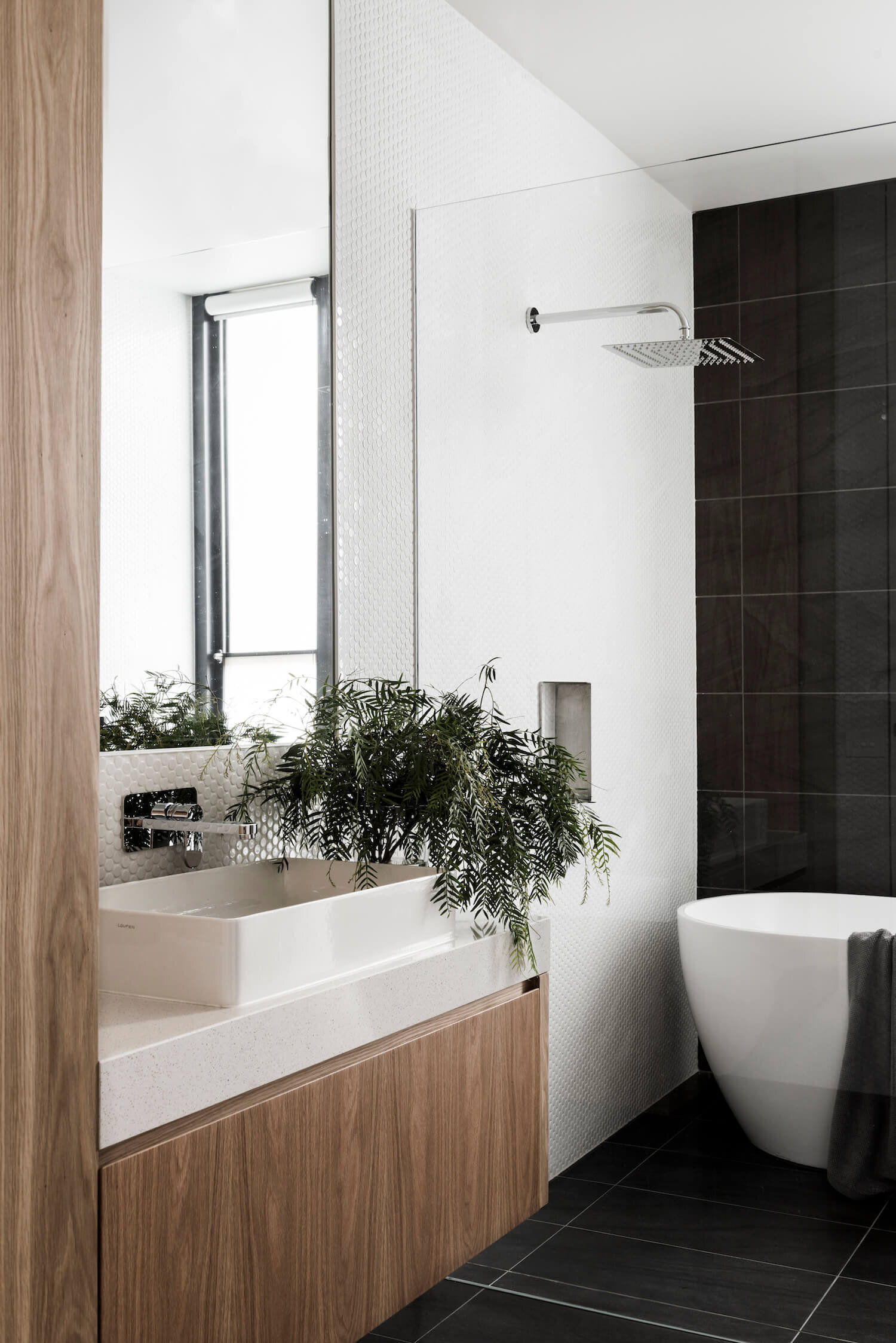 The bathroom takes on a bolder, contemporary tone. Large dark tiles are paired with smaller white ones, creating visual balance. Touches of wood soften the space, ensuring it remains warm rather than stark. The result is a bathroom that feels both sophisticated and inviting.