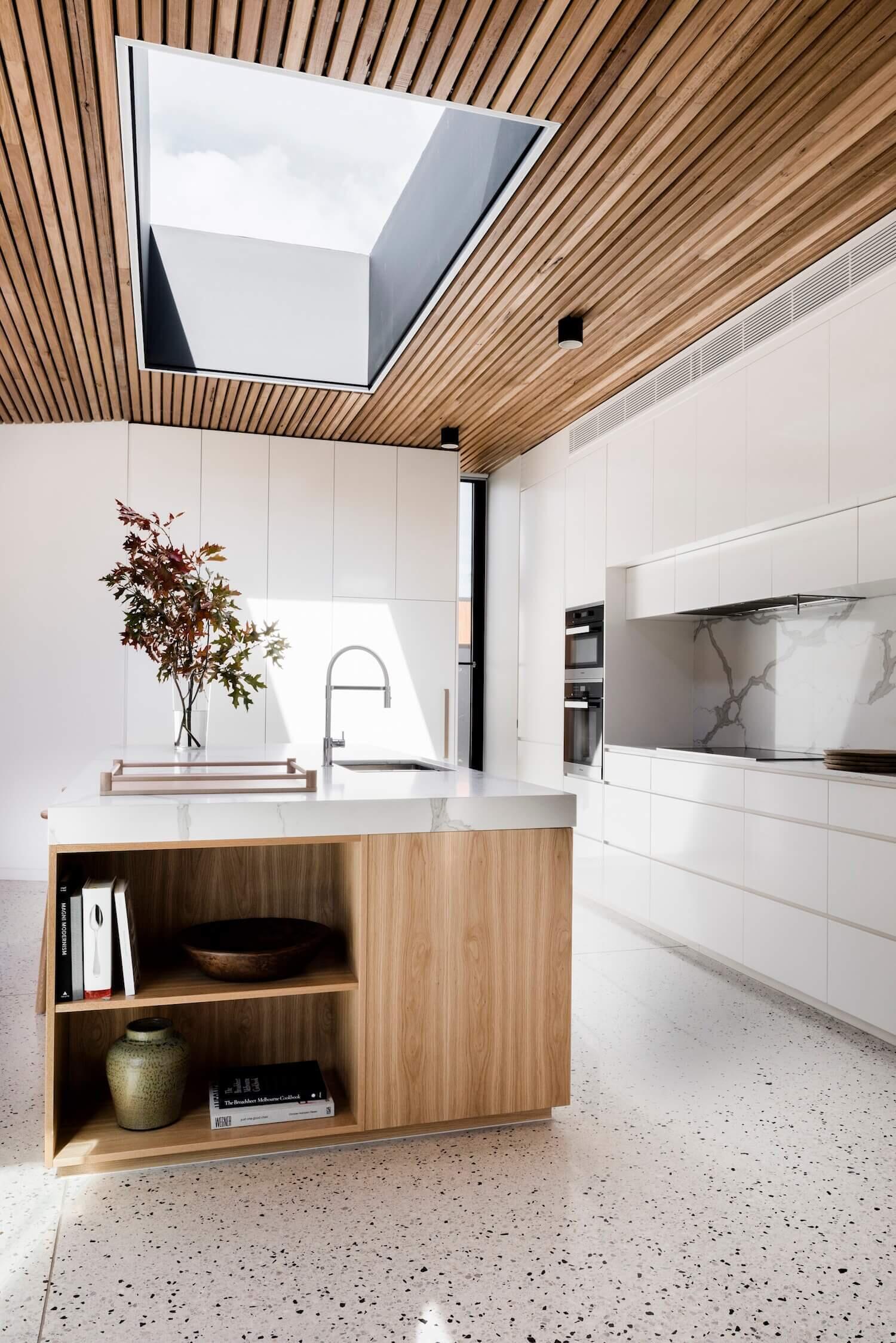 Opposite the living area sits the dining room and kitchen. A light wood dining table and chairs echo the timber ceiling and bring harmony to the palette. The kitchen itself is designed with a modern, understated elegance. White cabinets are paired with a stone backsplash and countertop, while the island introduces natural warmth in the form of light wood, complete with seating and open shelving.