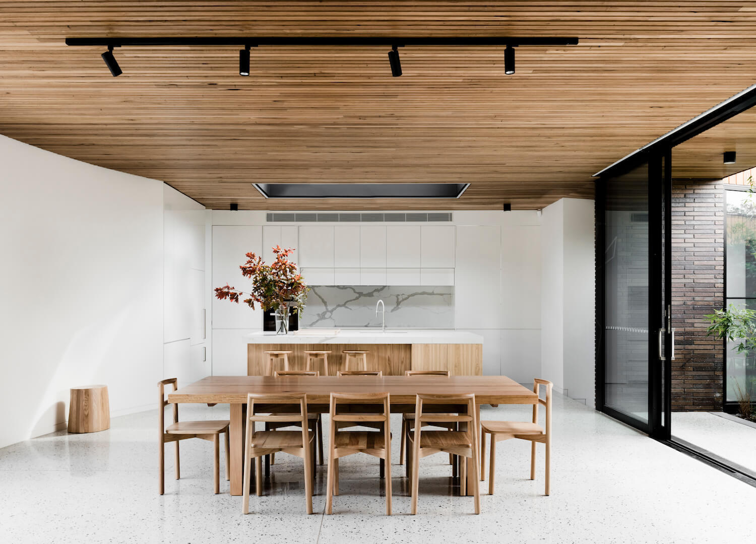Opposite the living area sits the dining room and kitchen. A light wood dining table and chairs echo the timber ceiling and bring harmony to the palette. The kitchen itself is designed with a modern, understated elegance. White cabinets are paired with a stone backsplash and countertop, while the island introduces natural warmth in the form of light wood, complete with seating and open shelving.