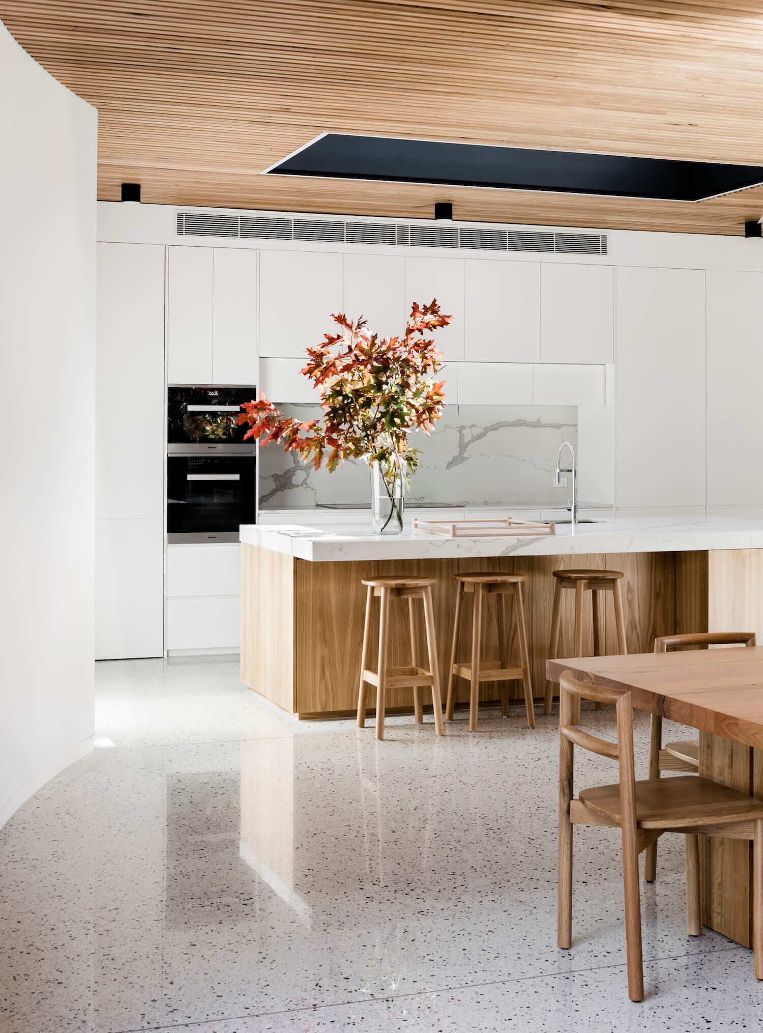 Opposite the living area sits the dining room and kitchen. A light wood dining table and chairs echo the timber ceiling and bring harmony to the palette. The kitchen itself is designed with a modern, understated elegance. White cabinets are paired with a stone backsplash and countertop, while the island introduces natural warmth in the form of light wood, complete with seating and open shelving.