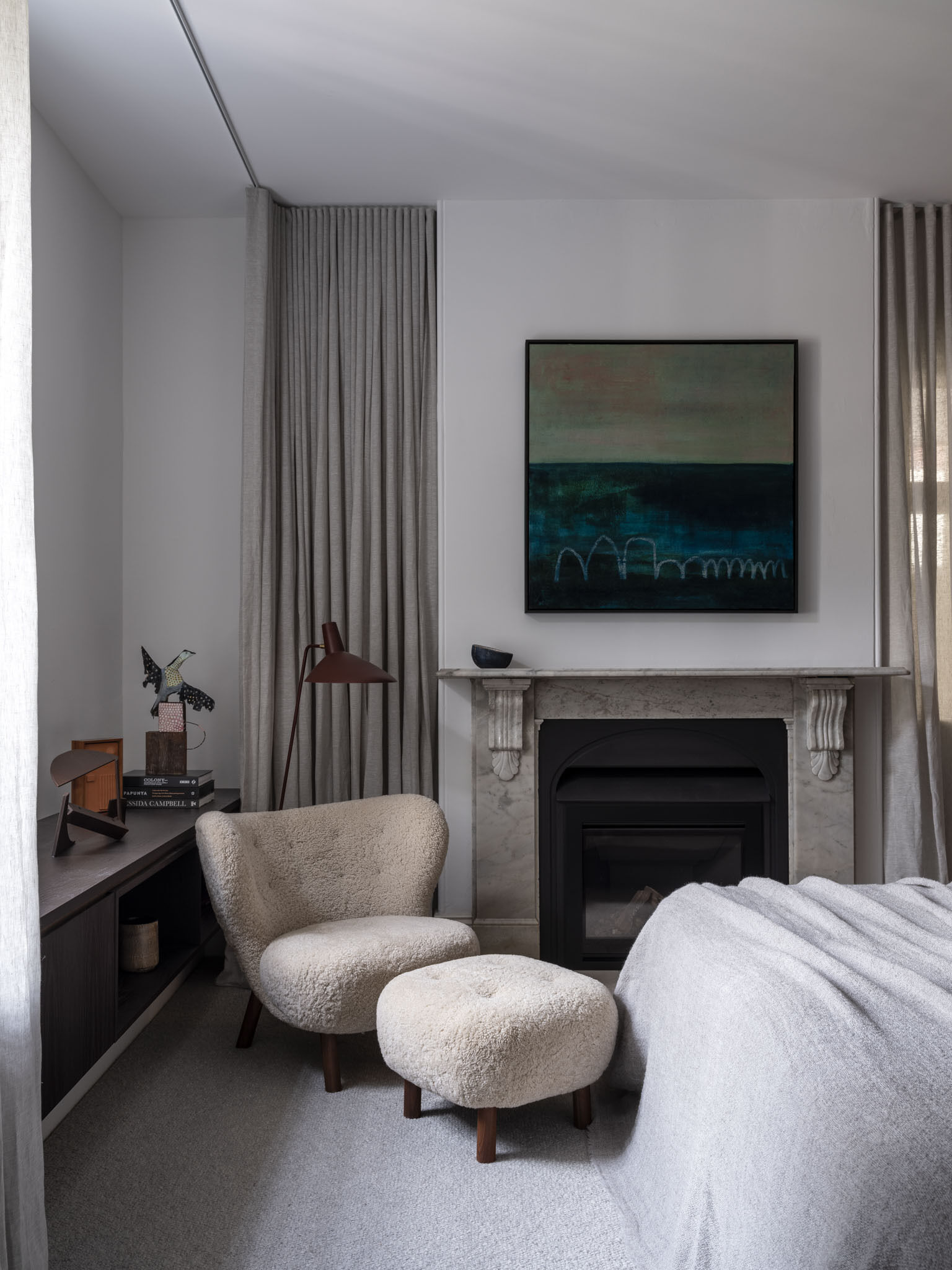 Soft daylight filters through the window in the primary bedroom, washing the room in a gentle glow that highlights its calm palette of whites and warm neutrals. A cozy boucle chair with matching ottoman sits by the fireplace and clean-lined built-ins. Layered details like the linen curtains, wool throw, and ceramic accents make the space feel effortlessly comfortable, while its deep charcoal walls and vertical wood paneling behind the bed add depth and rhythm.