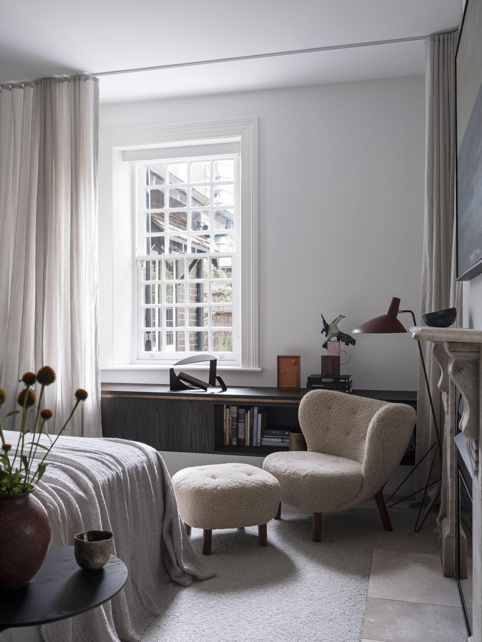 Soft daylight filters through the window in the primary bedroom, washing the room in a gentle glow that highlights its calm palette of whites and warm neutrals. A cozy boucle chair with matching ottoman sits by the fireplace and clean-lined built-ins. Layered details like the linen curtains, wool throw, and ceramic accents make the space feel effortlessly comfortable, while its deep charcoal walls and vertical wood paneling behind the bed add depth and rhythm.