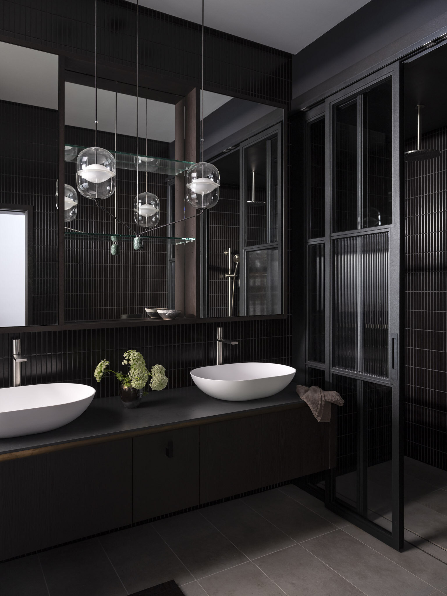 This ensuite bathroom pairs drama with calm through its mix of dark tiled walls and soft gray flooring. The double vessel sinks in crisp white stand out like sculptural accents, while the glass globe pendants add a touch of elegance and gentle light.