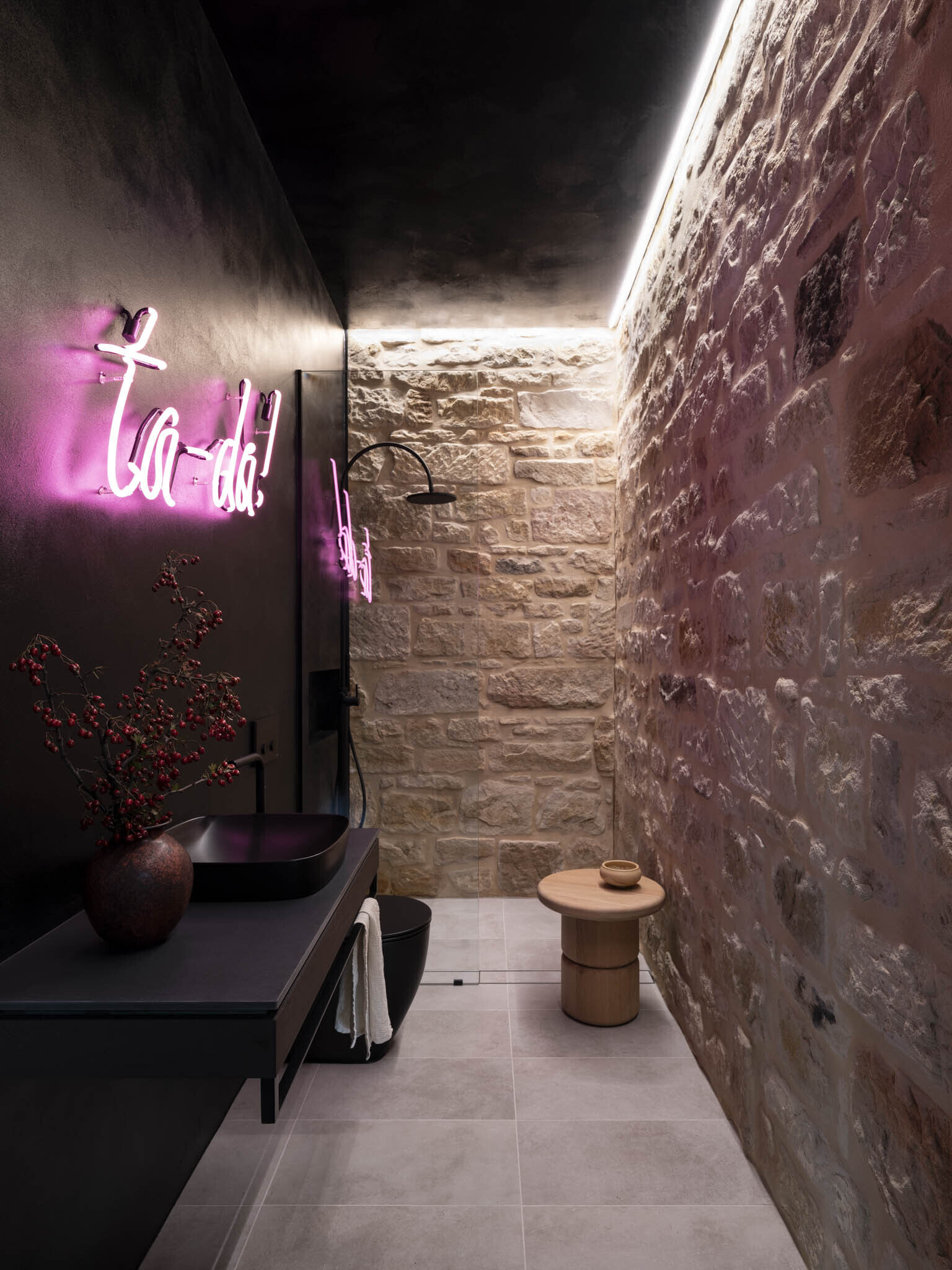 This shower room feels like a playful hideaway, blending raw stone walls with sleek black finishes. The warm texture of the masonry contrasts beautifully with the smooth surfaces of the vanity and fixtures, while soft gray floor tiles ground the space, and a neon pink sign adds unexpected fun, its glow bouncing off the stone for a lively touch.