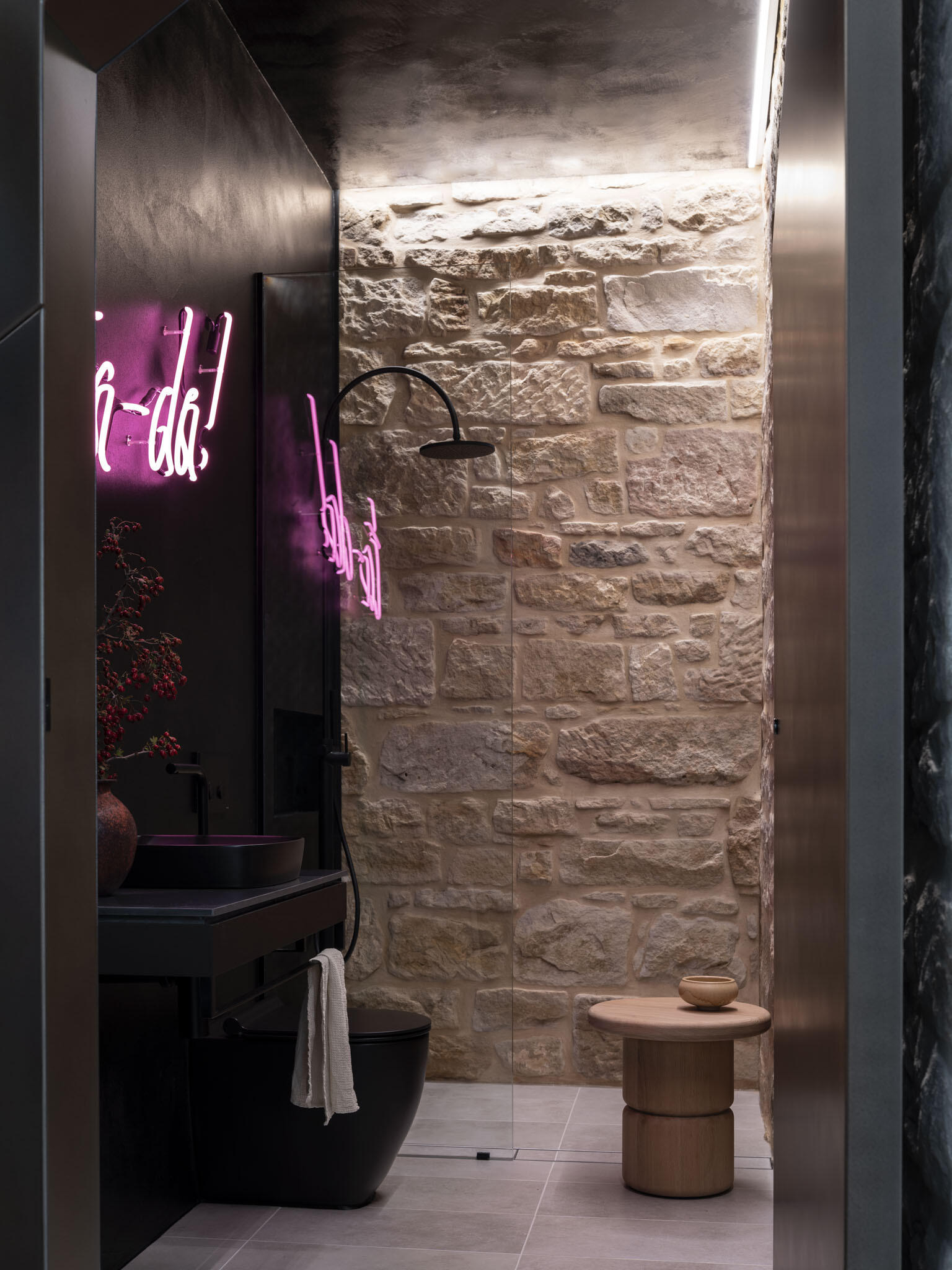 This shower room feels like a playful hideaway, blending raw stone walls with sleek black finishes. The warm texture of the masonry contrasts beautifully with the smooth surfaces of the vanity and fixtures, while soft gray floor tiles ground the space, and a neon pink sign adds unexpected fun, its glow bouncing off the stone for a lively touch.