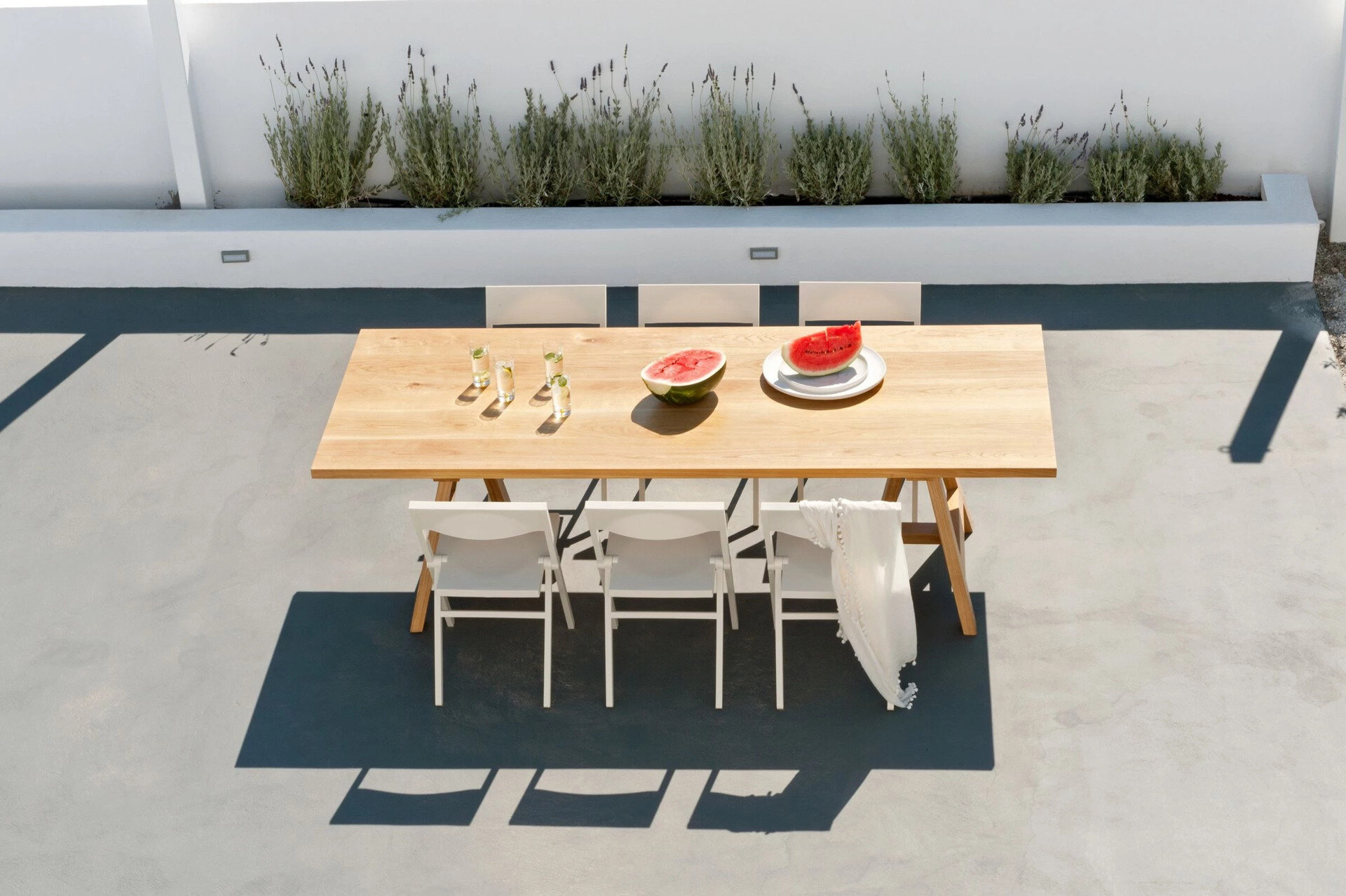 A long wooden table, white chairs, and a sunlit garden define the home’s outdoor dining area, the perfect setting for summer evenings on the island.