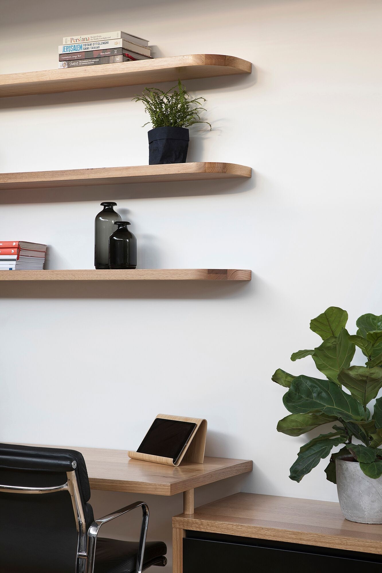 The home office desk runs seamlessly into the living area, transforming into an entertainment cabinet. Above it, floating timber shelves with softly curved corners echo the geometry of the clerestory windows.