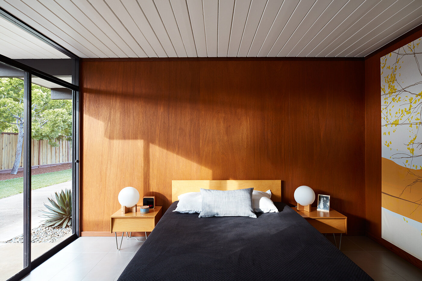 A serene mid-century bedroom connects to a private patio through sliding glass doors and a continuous wood ceiling.