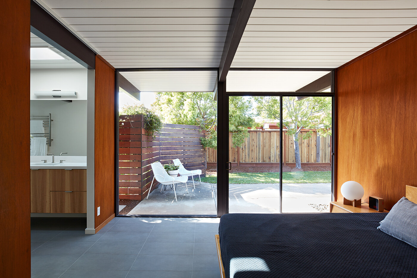 A serene mid-century bedroom connects to a private patio through sliding glass doors and a continuous wood ceiling.