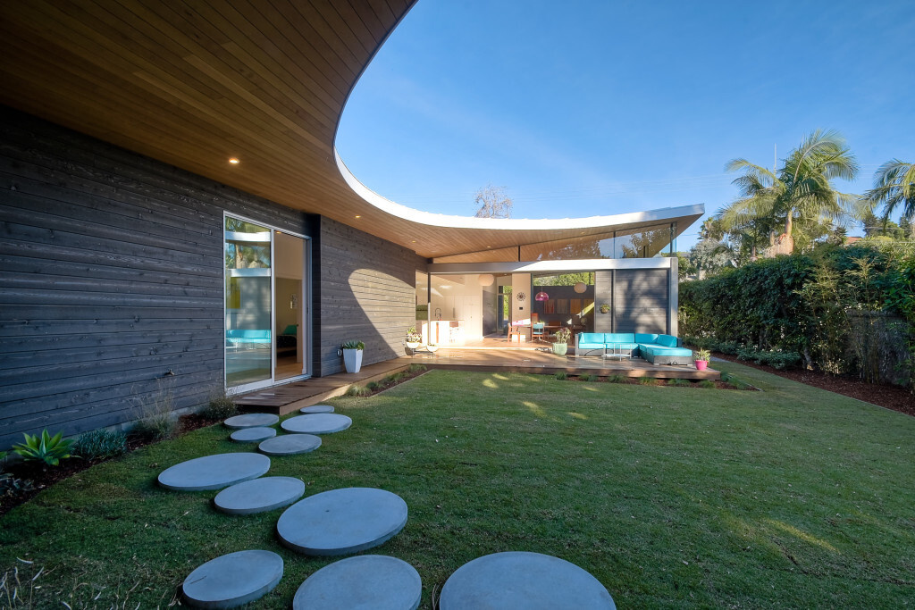 A sweeping curved roof defines the architecture of Avocado Acres House, uniting three pavilions around a central courtyard. Influenced by mid-century modern design yet built with 21st-century sustainability, the home blends bold geometry, natural light, and playful details for a striking coastal residence.