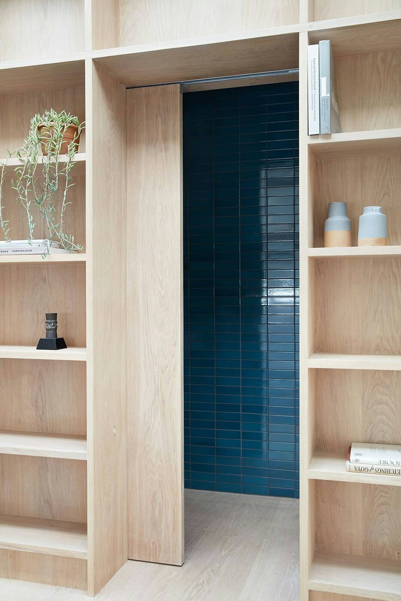 Tucked behind wooden shelving, this hidden powder room reveals bold blue tiles that bring unexpected energy to the refined studio.