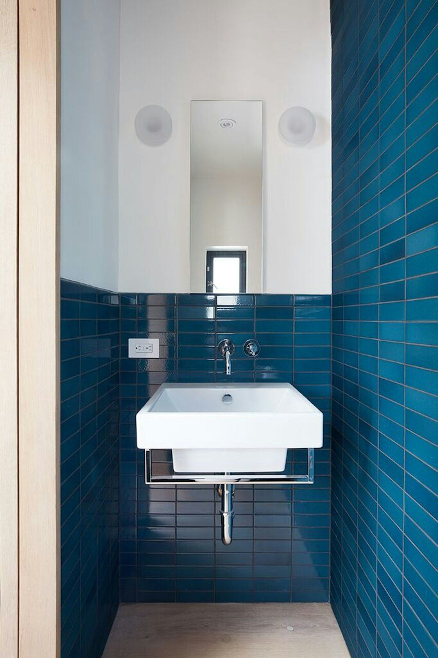 Tucked behind wooden shelving, this hidden powder room reveals bold blue tiles that bring unexpected energy to the refined studio.