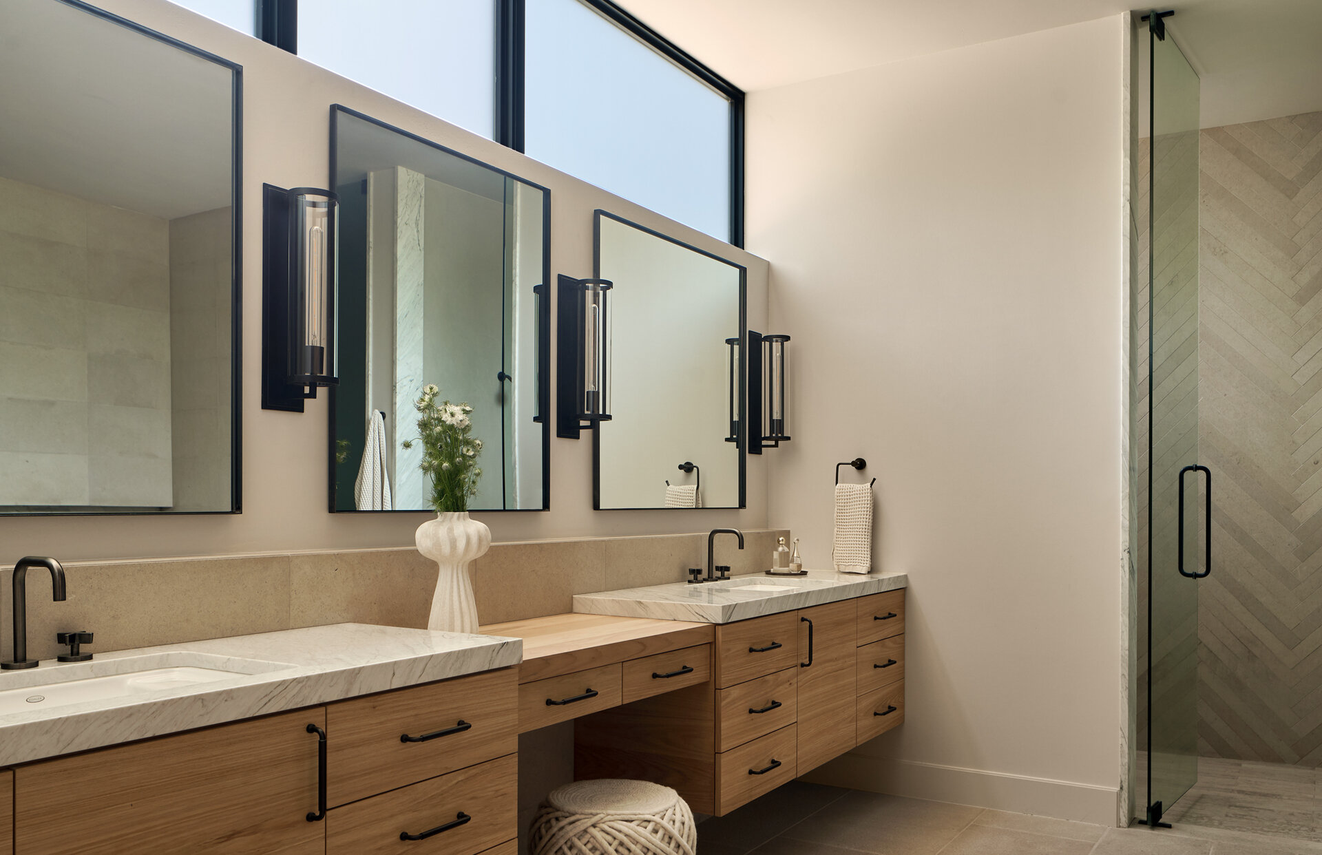 This bathroom combines calm elegance with thoughtful detailing. Dual sinks set in marble-topped vanities anchor the space, with warm wood cabinetry adding texture and storage. To one side, a glass-enclosed shower lined in herringbone stone completes the composition, while subtle touches, like a sculptural vase and woven stool, add warmth and personality.