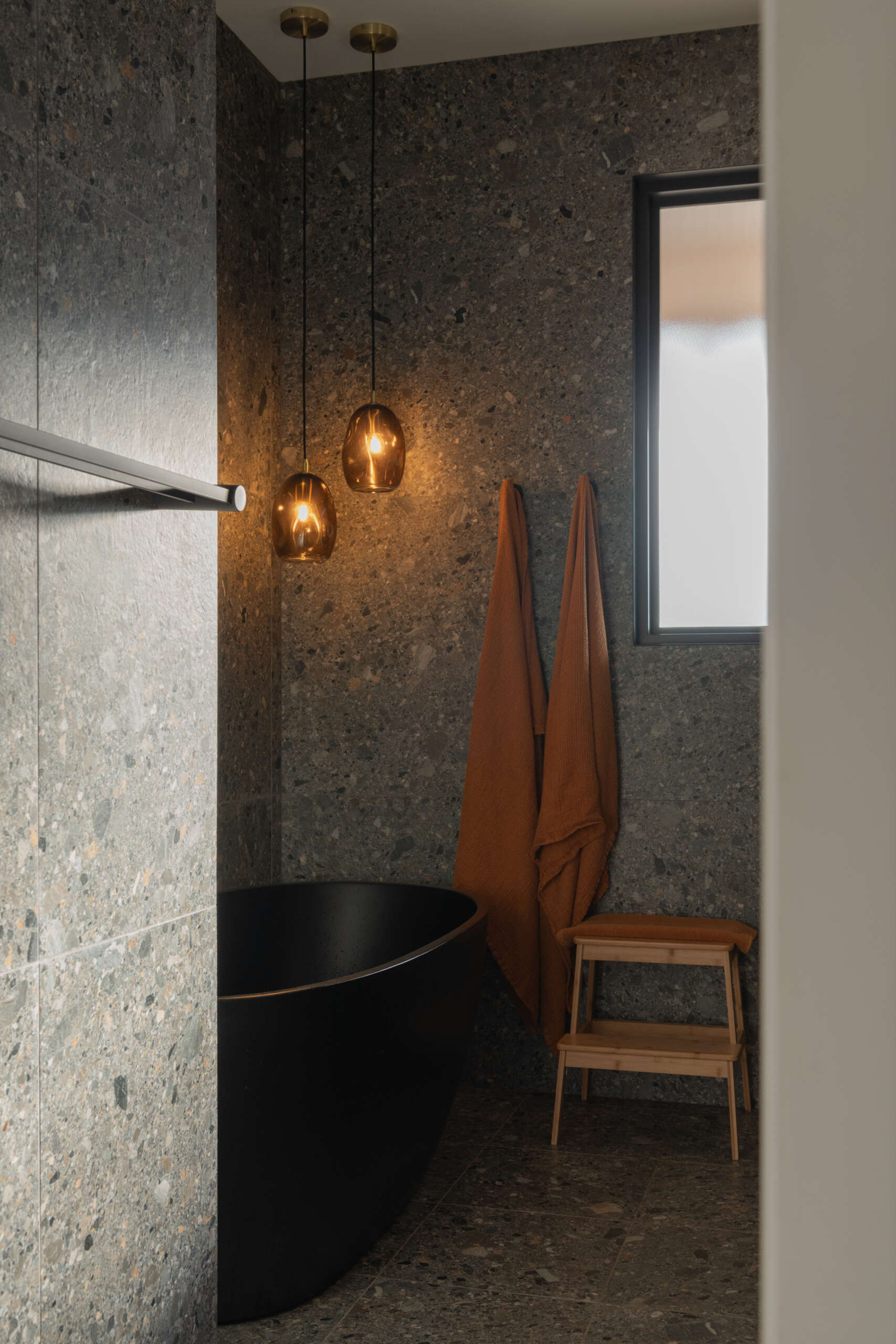 A mix of concrete, timber, and matte black fixtures defines the bathroom’s refined minimalism. The arched mirror and greenery introduce softness, transforming the space into a serene retreat.
